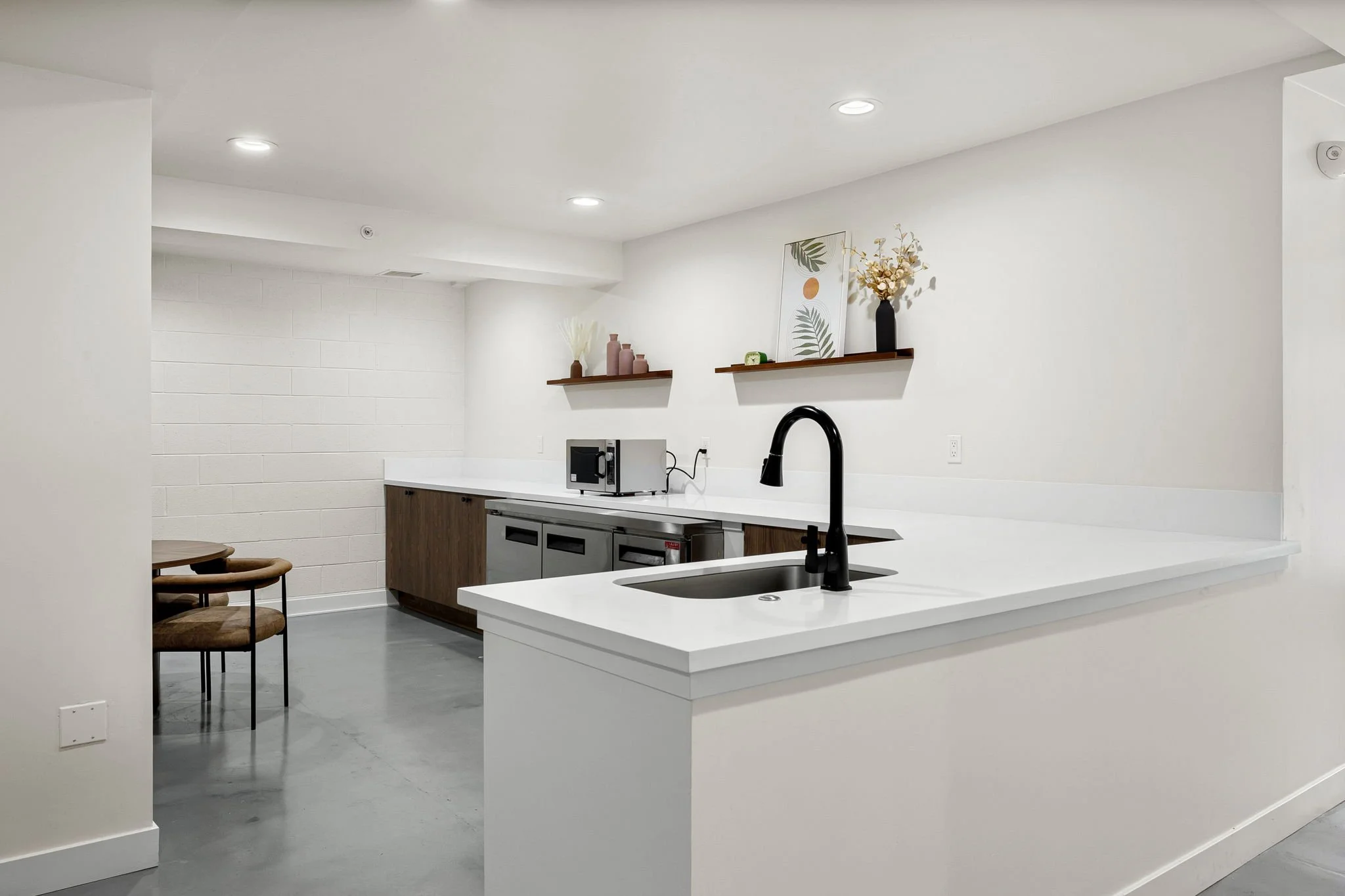 Modern kitchenette with white countertops, black faucet, brown cabinets, a microwave, and decorative shelves with vases and artwork.