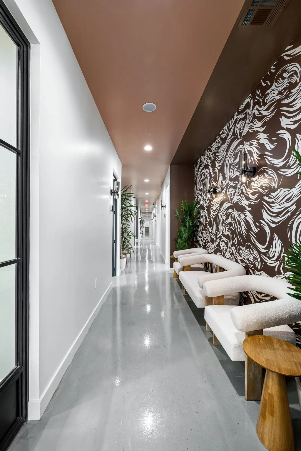Modern hallway with white walls, decorative wall art, green plants, and white chairs with wooden legs along the patterned wall.