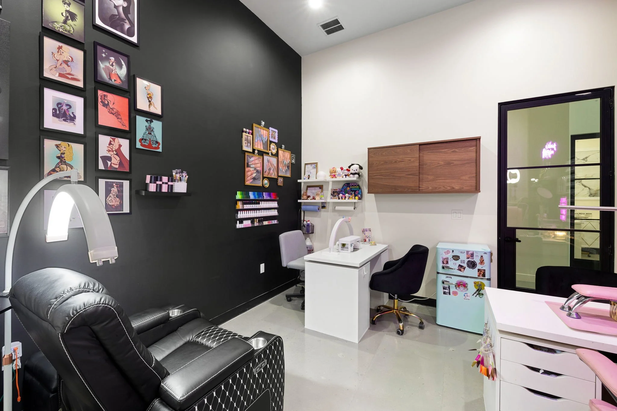 A beauty salon or spa treatment room with decor, framed artwork and nail polish display on a dark wall, a reclining massage chair, a manicure table with chairs, and a door leading outside.