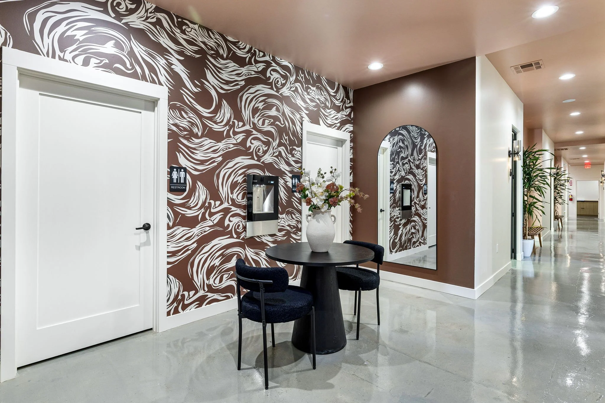 Modern interior hallway with brown and white swirling patterned accent wall, white restroom door, black round table with a white vase of flowers, black chairs, large mirror, and potted plants in the background.