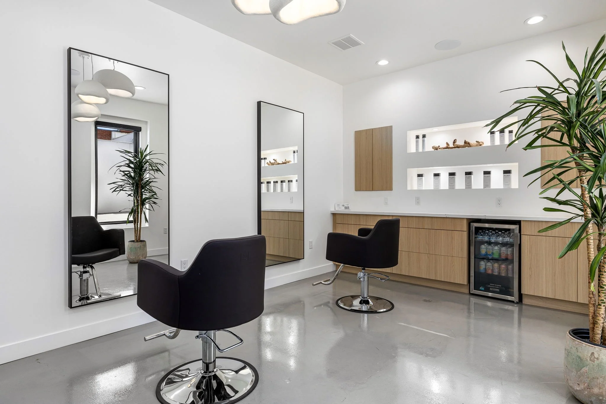 Modern hair salon with two black styling chairs, large mirrors, a potted plant, and a refrigerated beverage cooler.