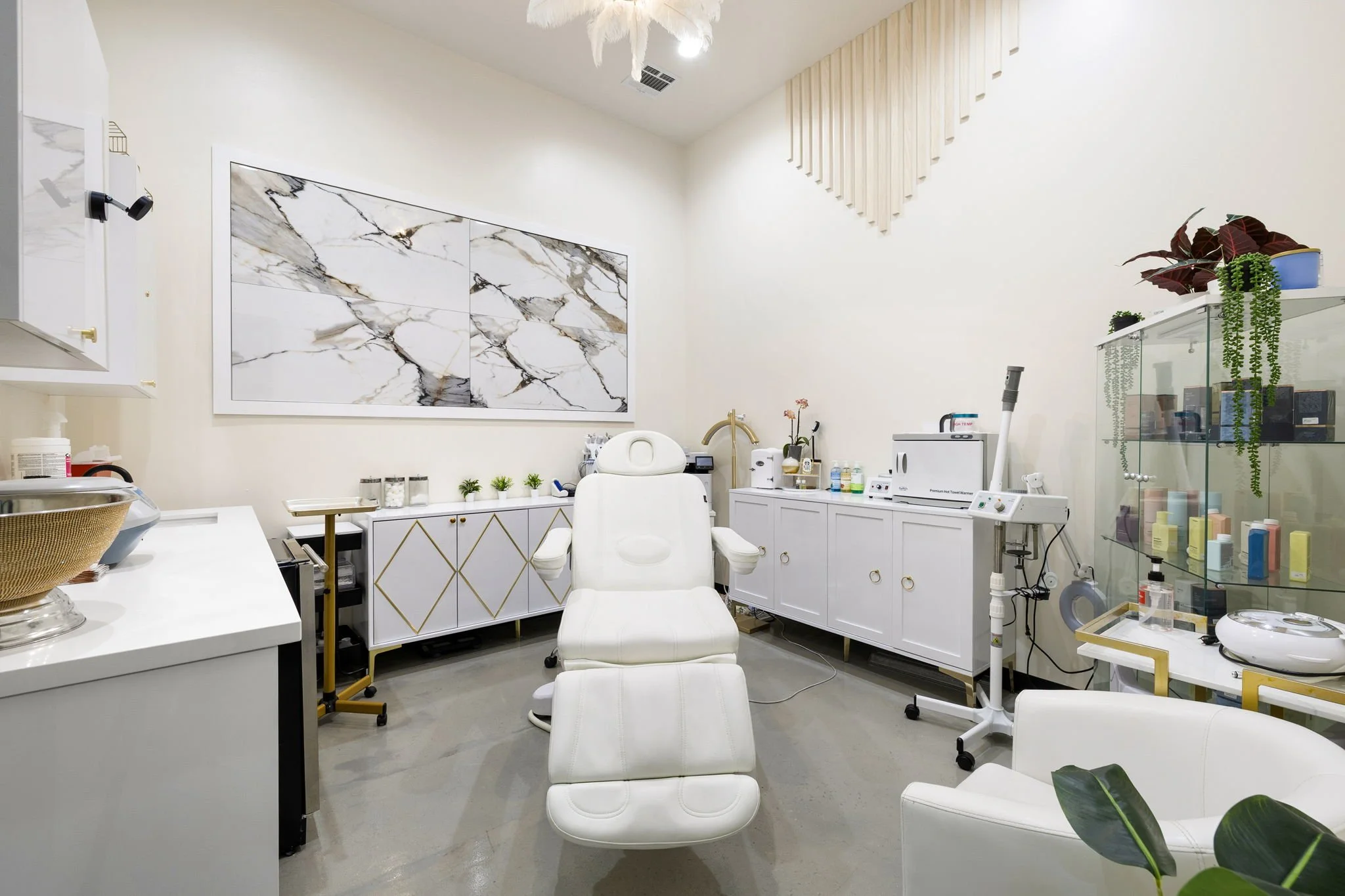 A modern aesthetic medical or dental treatment room with a white reclining chair centered, surrounded by white cabinetry, a marble wall art piece, and various medical equipment and supplies.