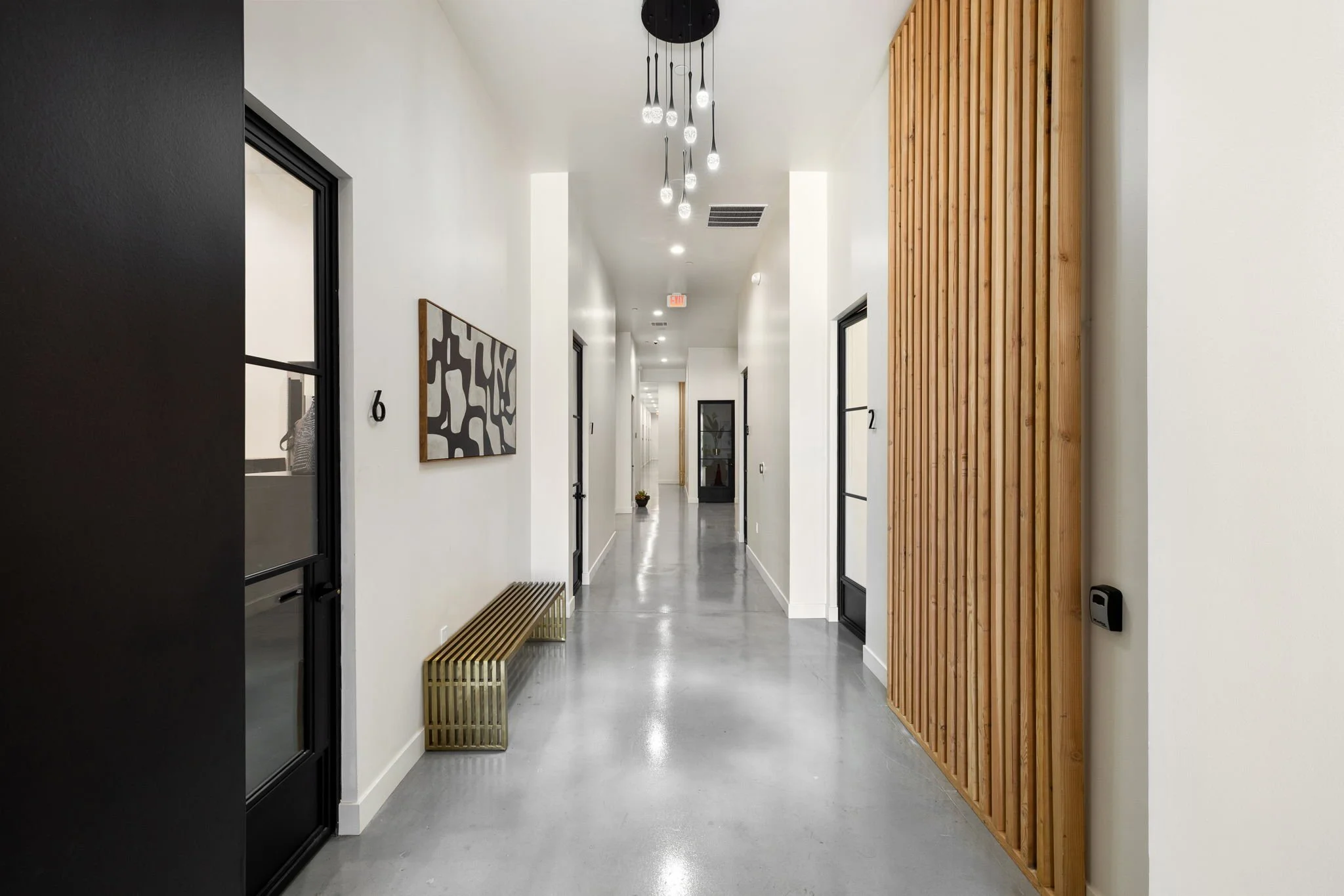 Modern hallway with white walls, black doors, a gold bench, a wooden slat wall, and a contemporary light fixture with hanging bulbs.
