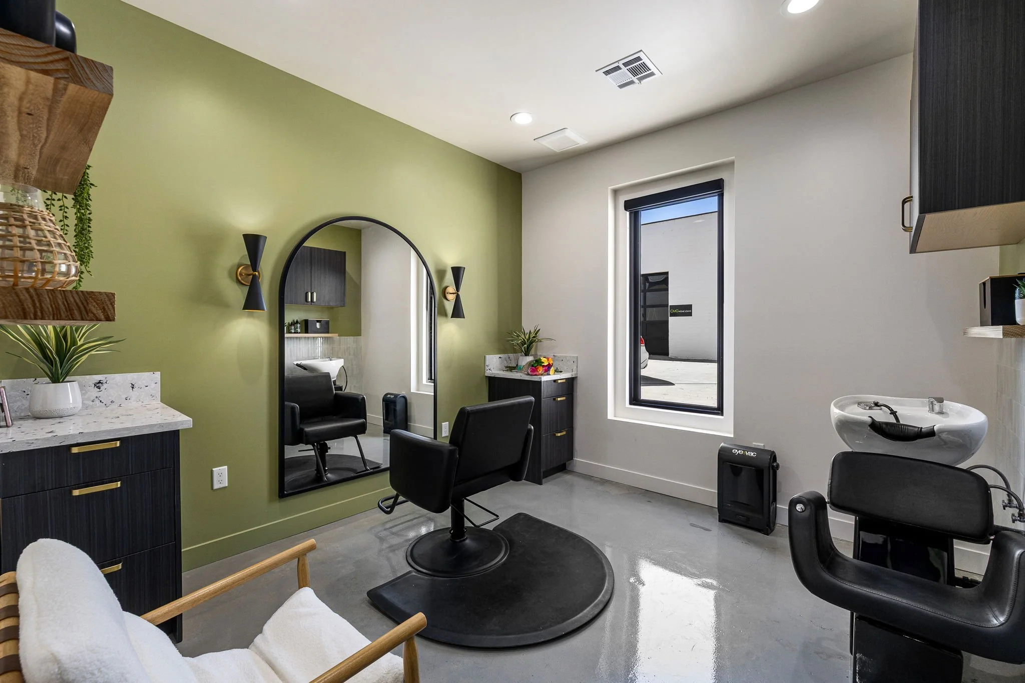 Modern barber or salon receiving area with black styling chairs, mirror, and hair washing station, decorated with plants and black, green, and white color scheme.