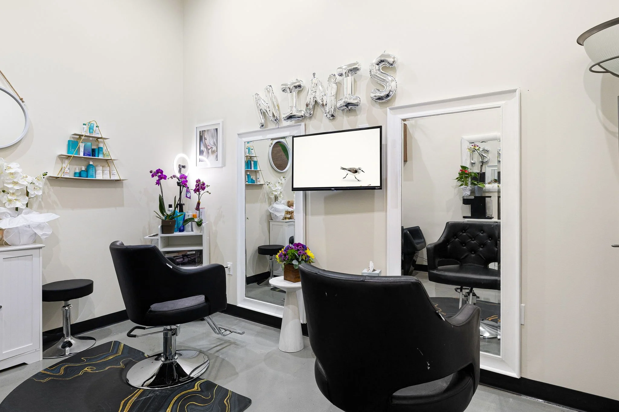 A nail salon with two black chairs, a small white table with flowers, mirrors, shelves with nail products, and balloon letters spelling 'NAILS' on the wall.
