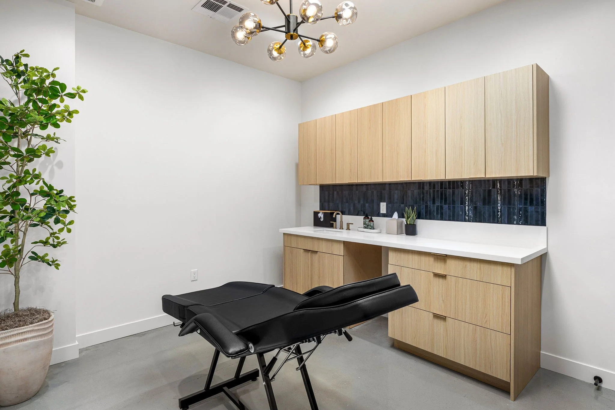 A modern treatment room with a black massage table, wooden cabinets, a white countertop, and a black tiled backsplash. A potted plant is on the left, and a contemporary chandelier hangs from the ceiling.