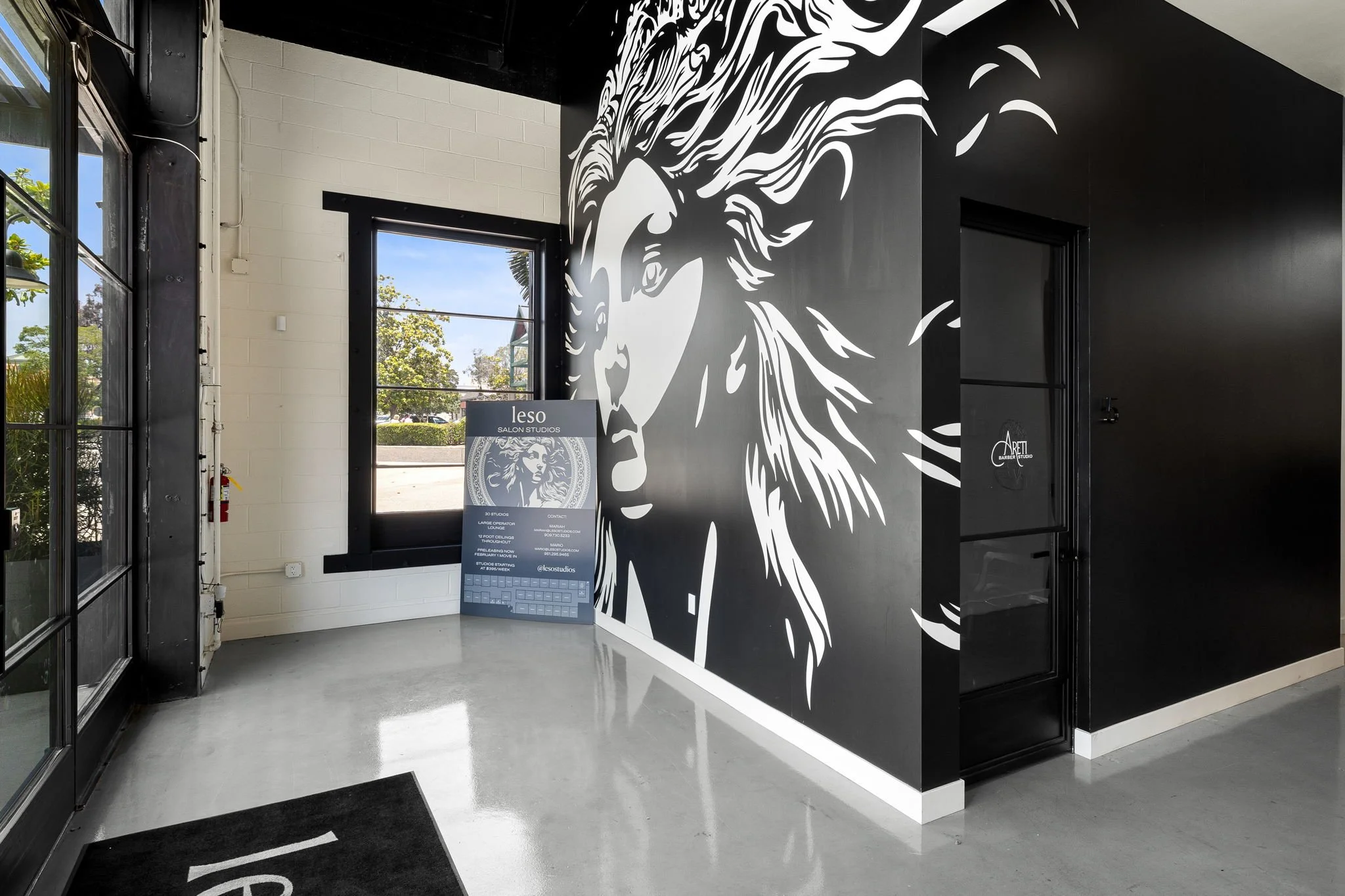 Interior of a modern salon or studio with a large black and white wall mural of a woman's face with flowing hair, black framed windows, and a standee sign near the window with salon information.