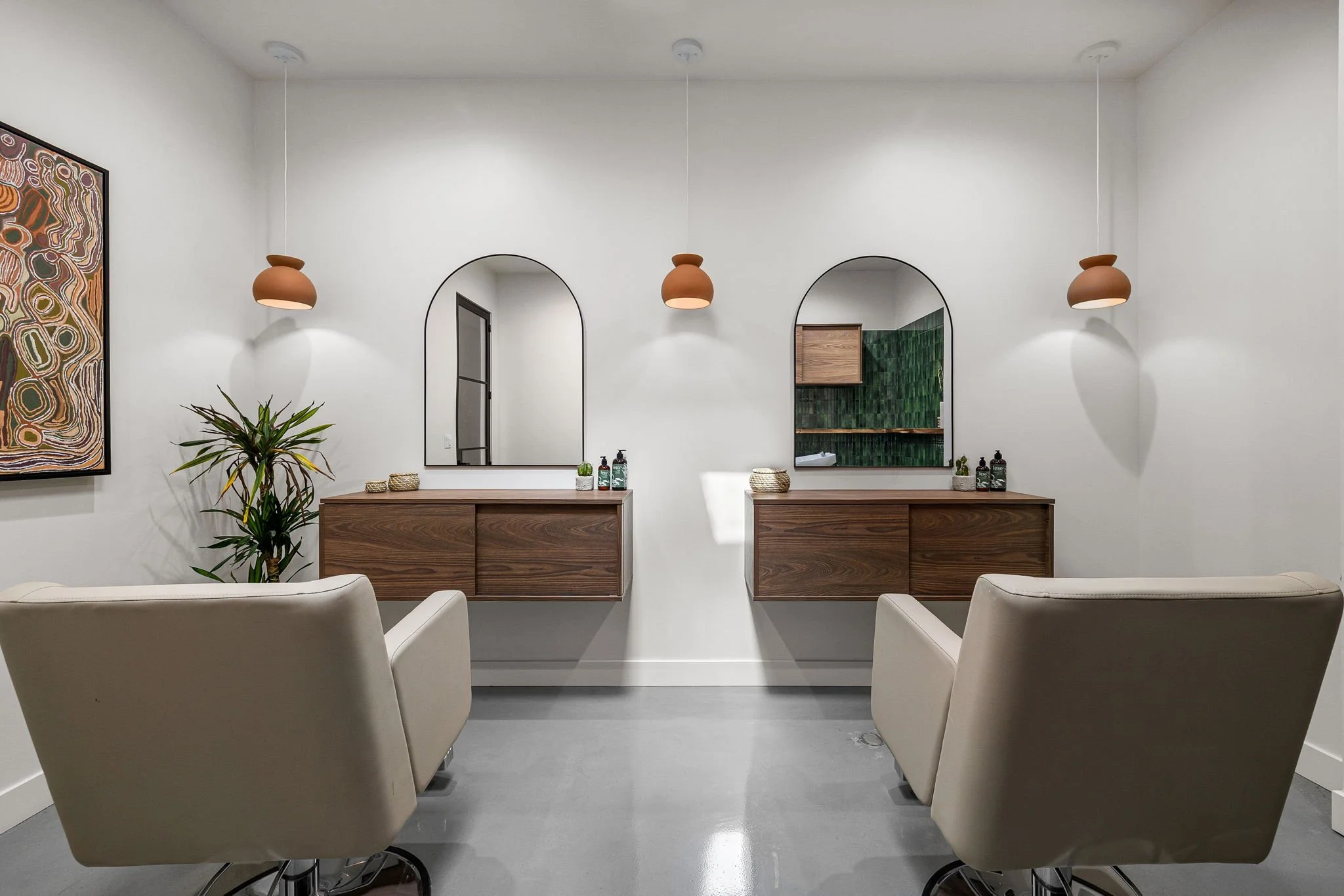 Minimalist hair salon or barber shop interior with two white chairs facing two wooden vanities with mirrors, decorative potted plants, and hanging pendant lights.