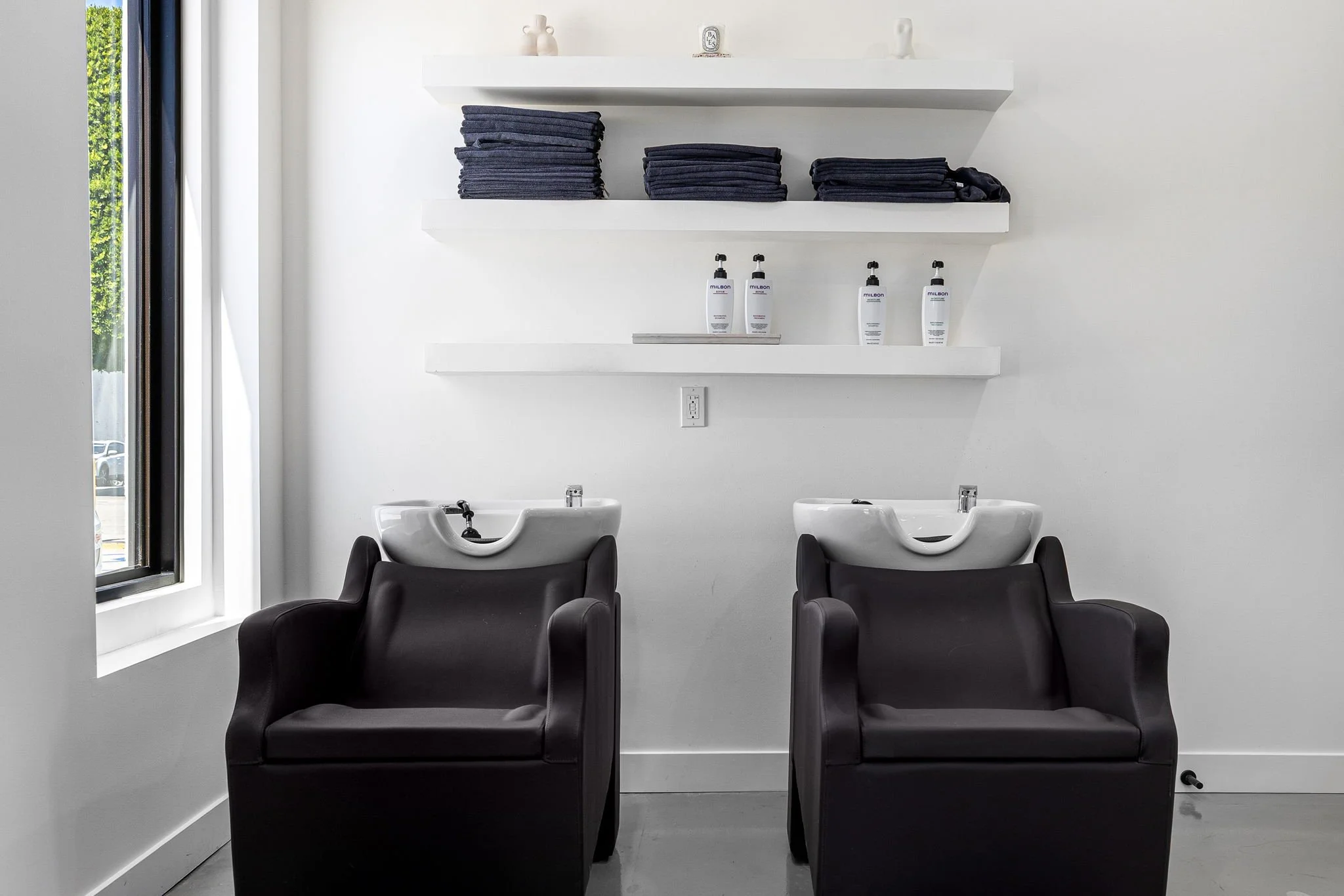 Two black salon chairs with white hair washing sinks behind them in a minimalist salon interior, with white walls, white shelves with folded towels and hair products, a large window on the left, and a Power outlet between the sinks.