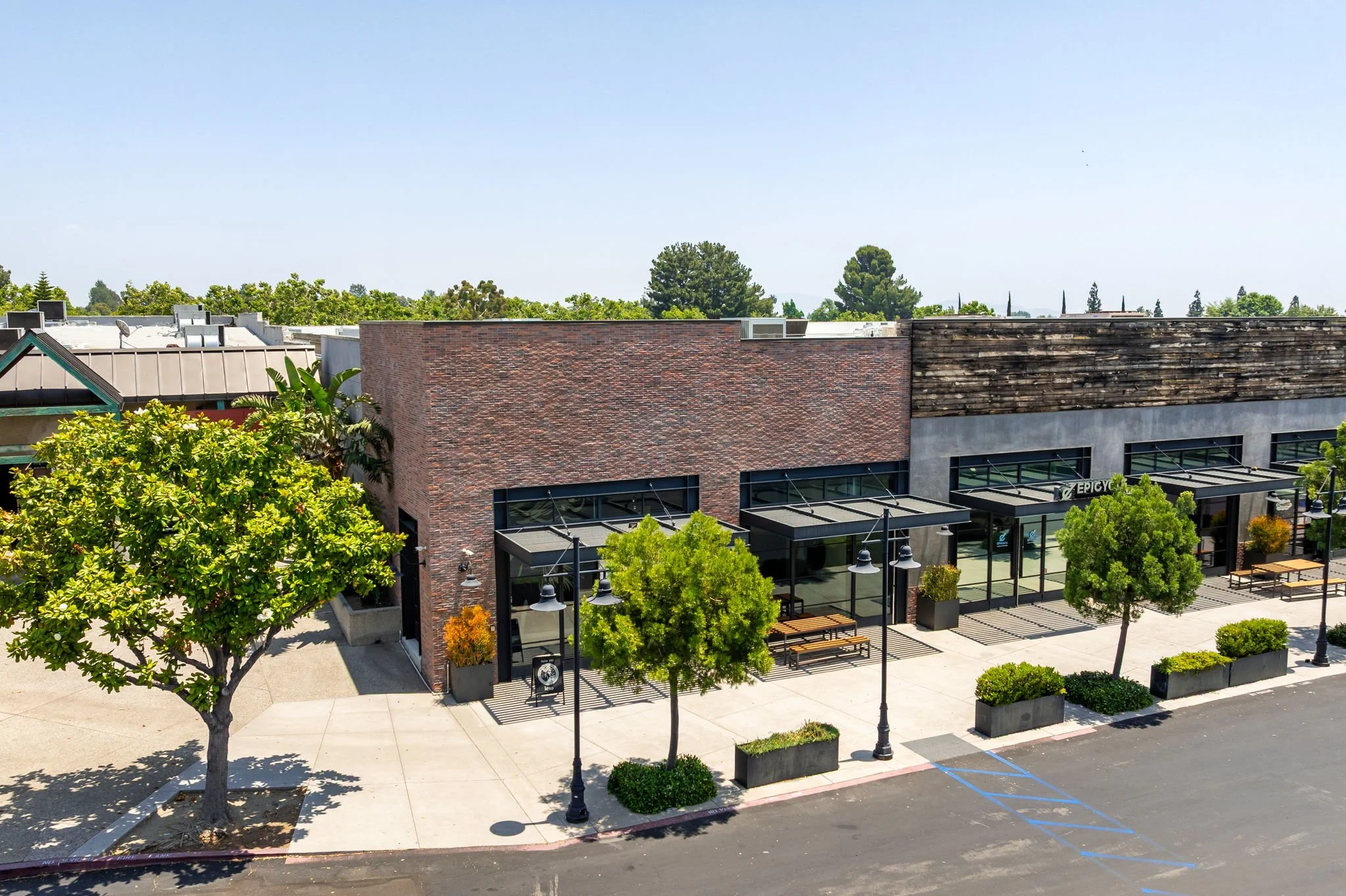 An outdoor shopping area with retail stores, trees, sidewalk, street, benches, and streetlights.