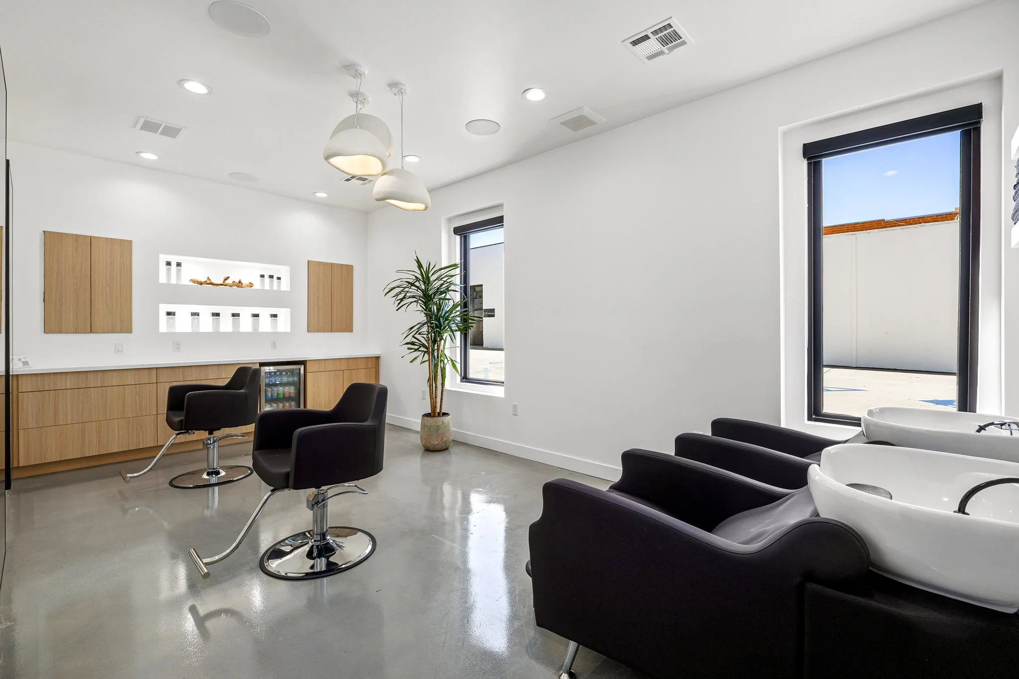 Inside a modern hair salon with two black hair washing sinks, three black salon chairs, and a wooden cabinet. There is a large window and a potted plant, with minimalistic decor and white walls.