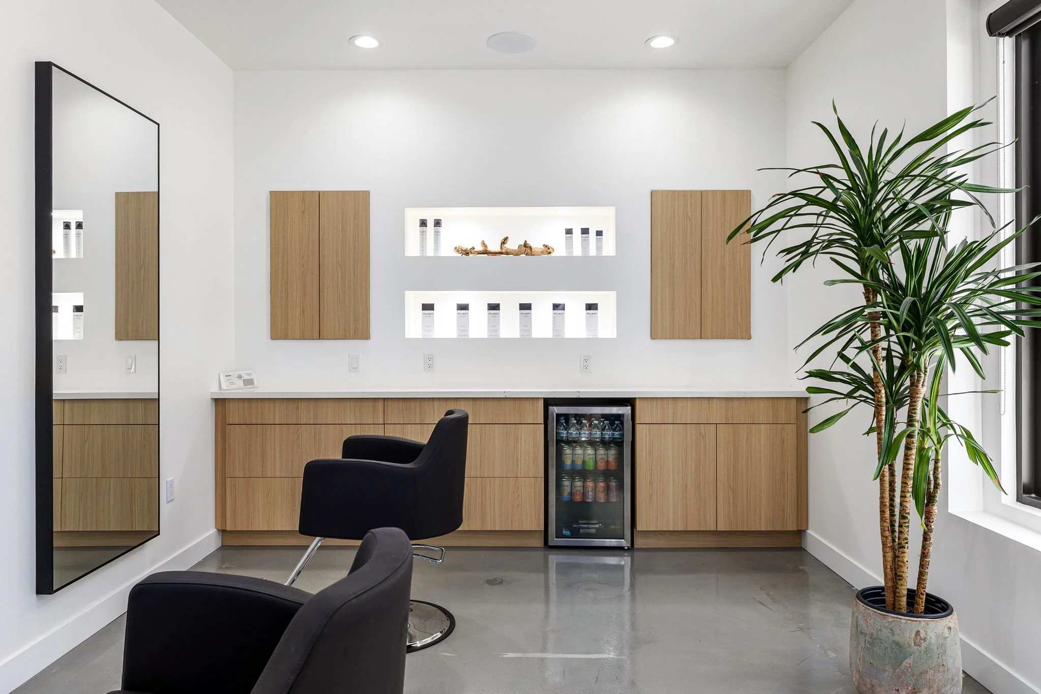 Modern hair salon or beauty studio with black chairs, large wall mirror, wooden cabinets, decorative shelves with products, mini refrigerator, and large potted plant by window.
