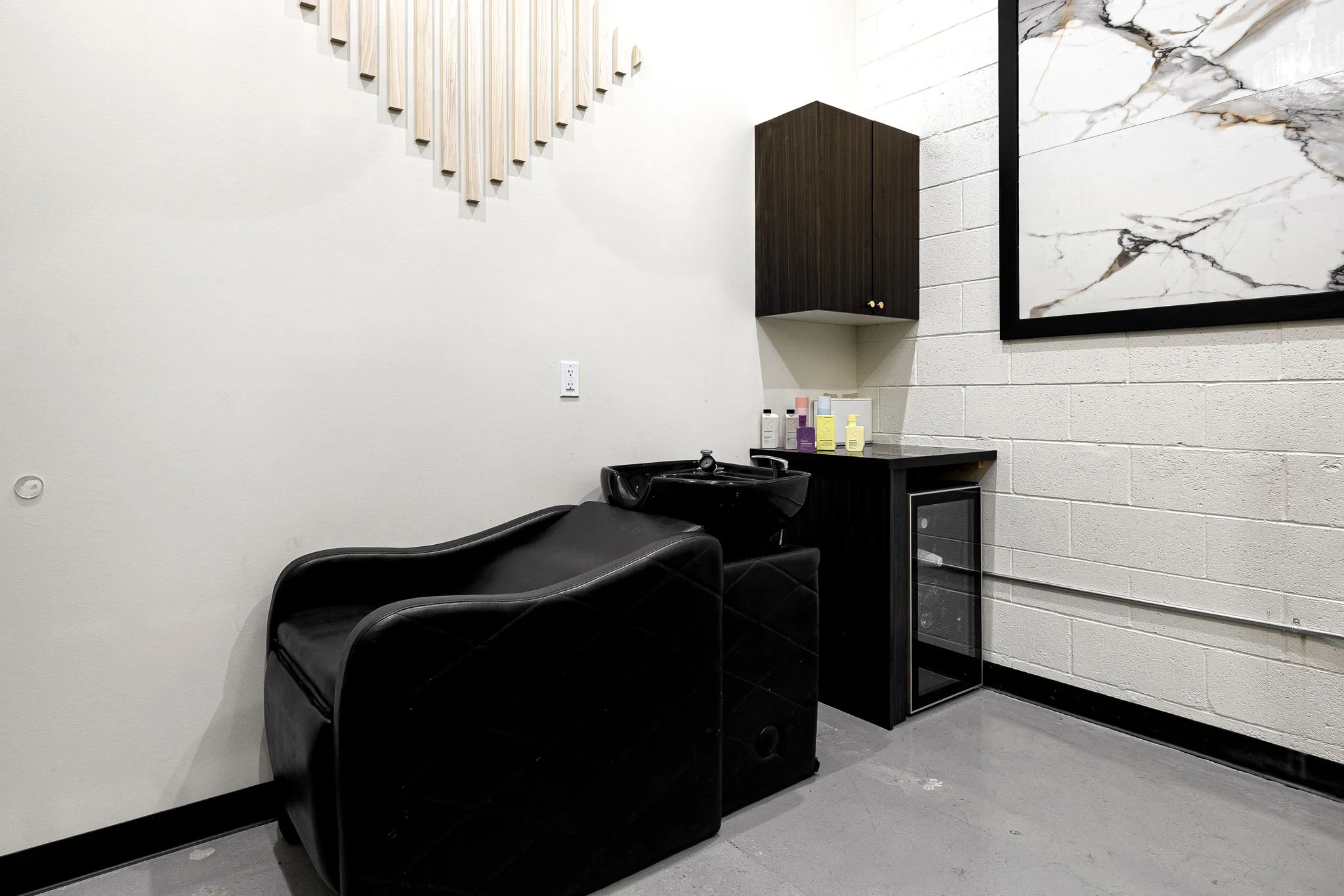 A salon shampoo station with a black reclining chair, black sink, small black cabinet, and a white wall with framed artwork.