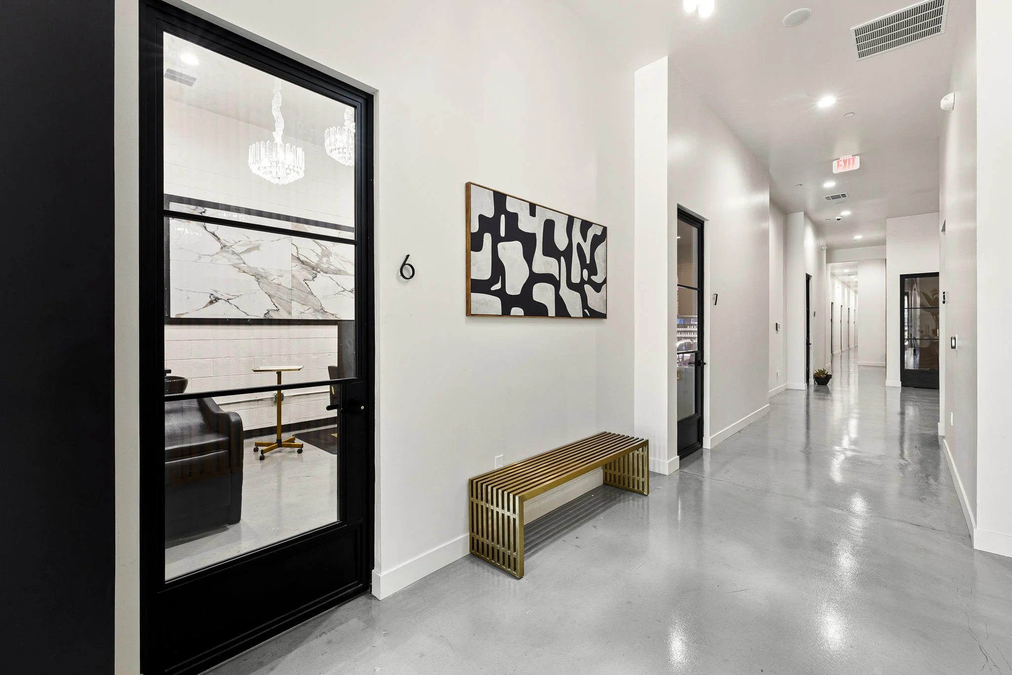Modern apartment hallway with white walls, gray polished concrete floors, a black door with glass, a gold art piece, a gold slatted bench, and a black picture frame.