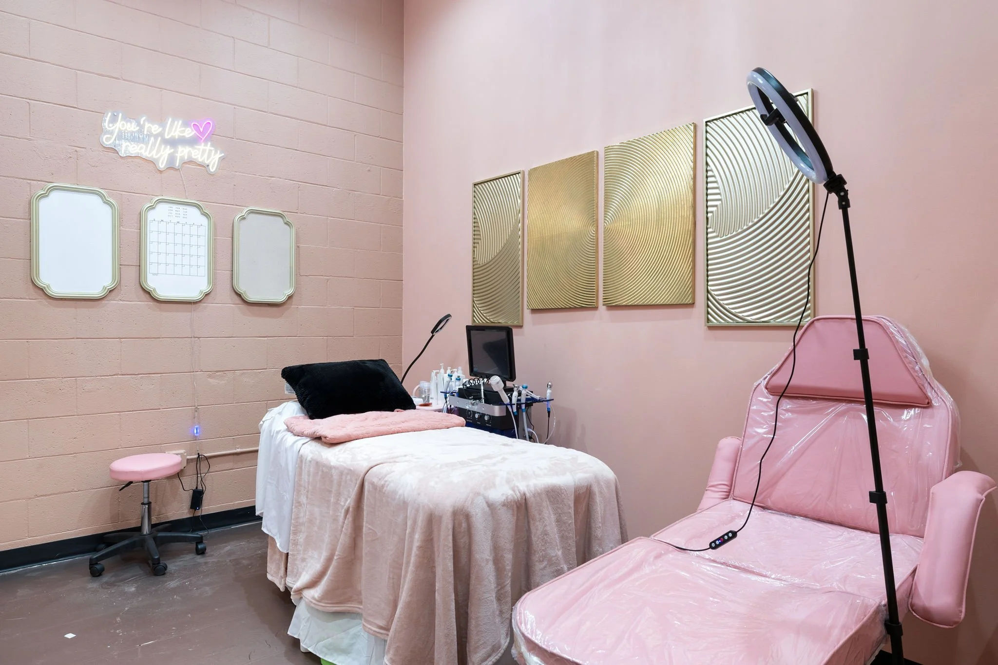 A pink-themed aesthetic room with a bed, beauty equipment, mirrors, and abstract wall art, suggesting a beauty or skincare treatment space.