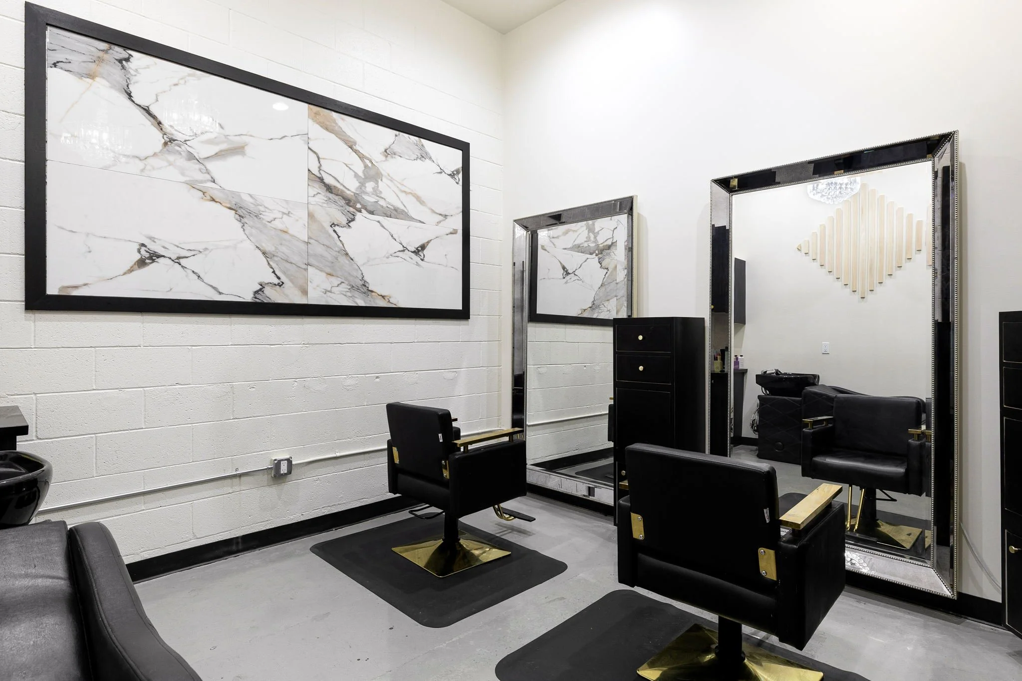 Interior of a modern hair salon with black and gold styling chairs, large marble wall art, and a tall decorative mirror with a glittering frame.