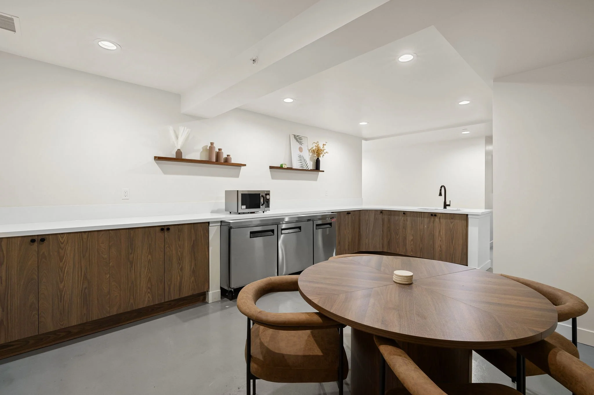 Modern kitchen with white walls, wooden cabinets, stainless steel appliances, a round wooden dining table with four chairs, and minimalist decor including shelves with vases and framed art.