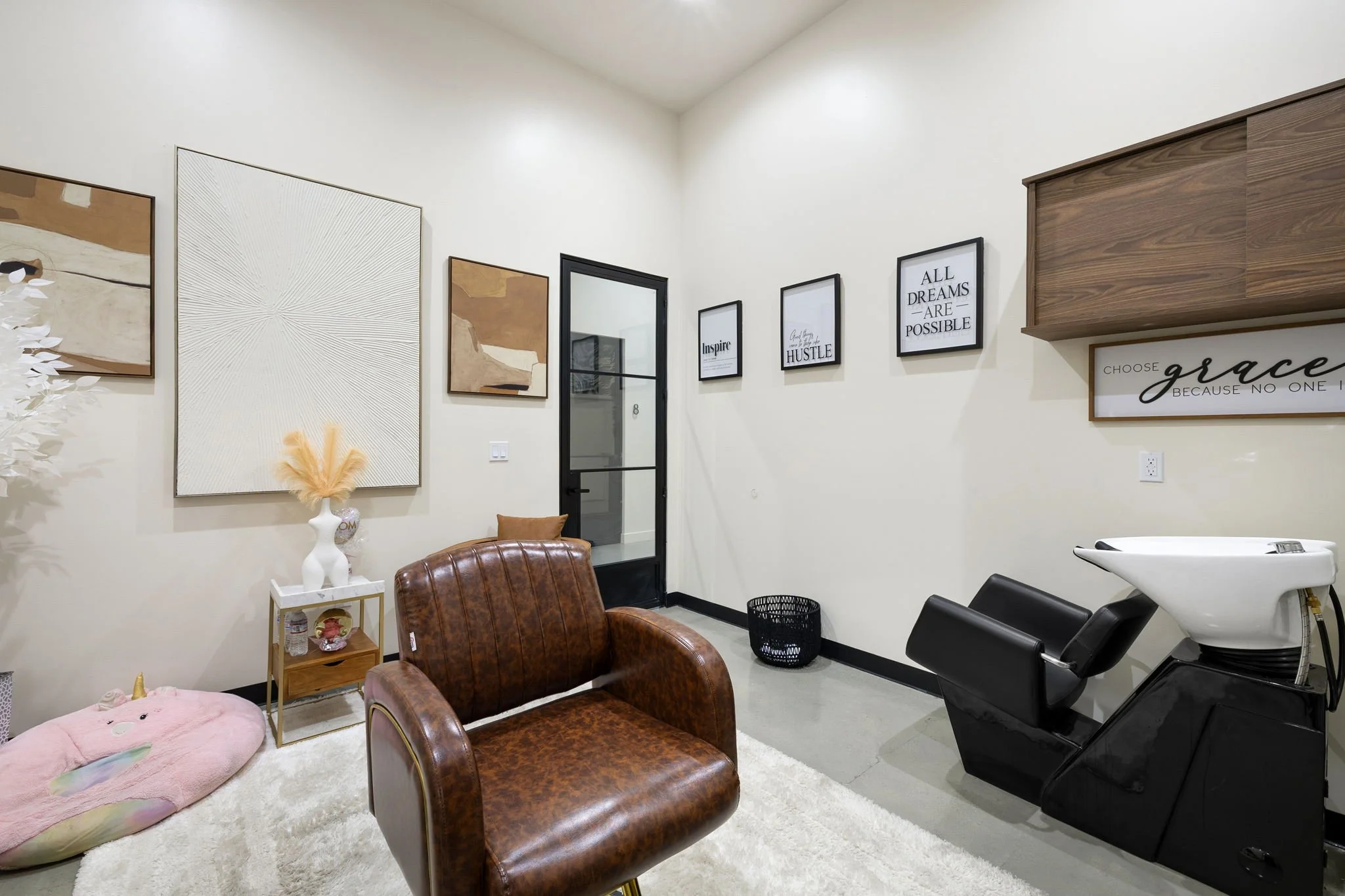 A modern hair salon with a brown leather armchair, a pink unicorn-shaped pet bed on the white rug, framed motivational posters on the wall, a small side table with decor and a water bottle, and a black shampoo station on the right side.