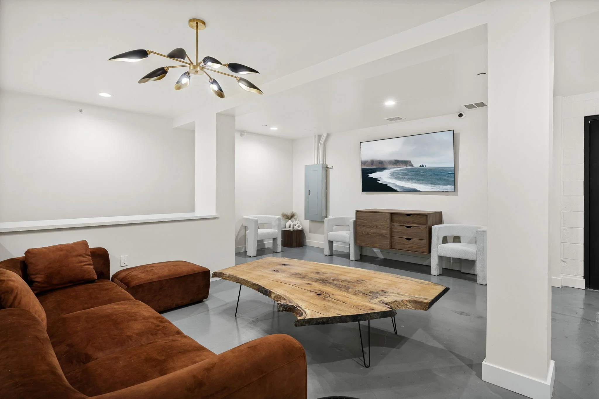 Living room with a brown velvet sectional sofa, a natural wood coffee table with metal hairpin legs, a wall-mounted flat-screen TV showing a coast landscape, a wooden console, white armchairs, and a modern black and gold chandelier.
