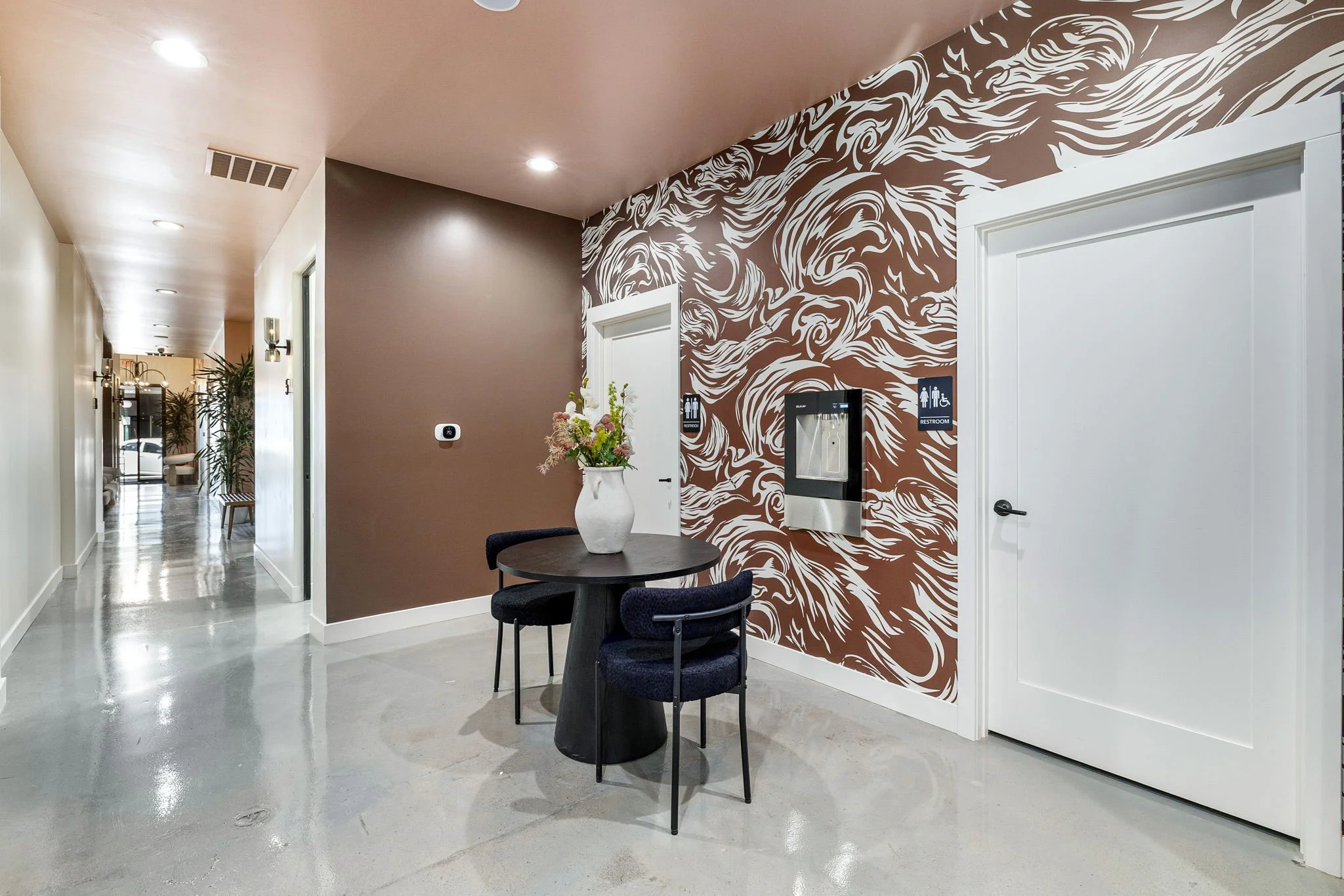 Modern interior with a small round table, two black chairs, a vase with flowers, and a decorative brown and white swirled wall. Restroom signs are visible on the wall.