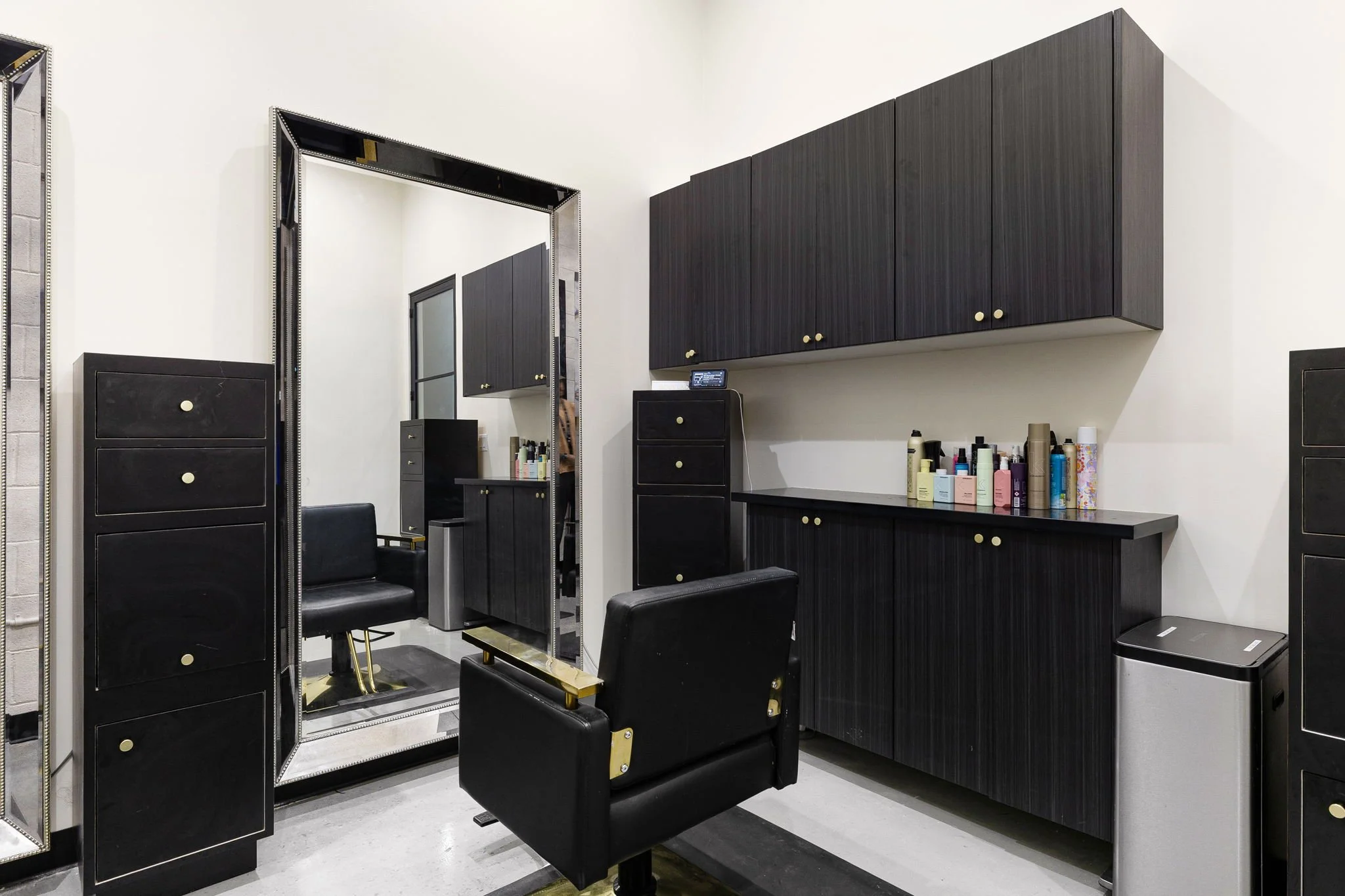 Hair salon station with large mirror, black swivel chair, black storage cabinets, and hair care products on the counter.