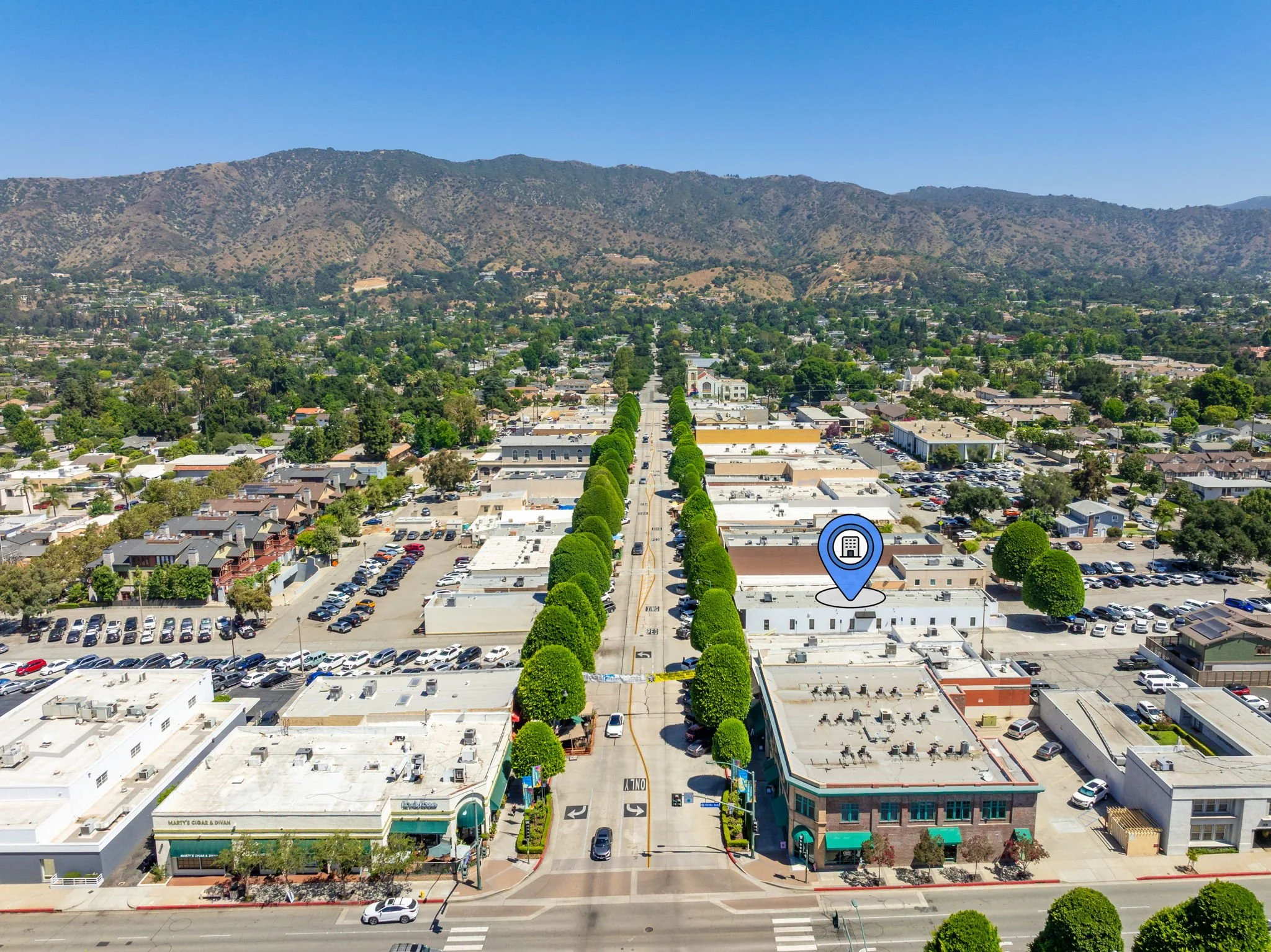Aerial view of a downtown area with a tree-lined street, commercial buildings, and parking lots, set against a backdrop of green hills and mountains under a clear blue sky.