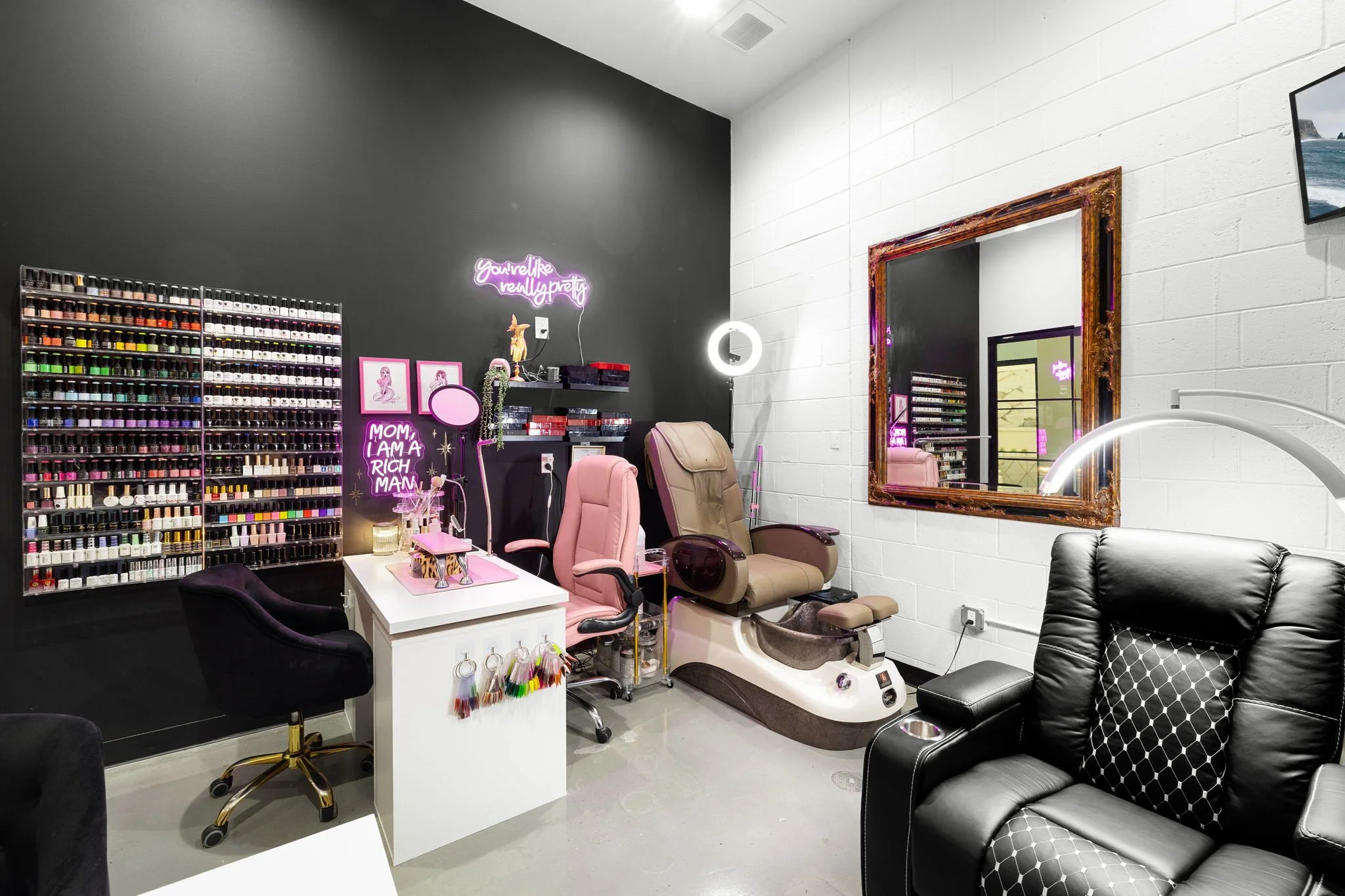 Nail salon with nail polish display, pink and black decor, massage chair, beauty-themed neon signs, mirror, and tinted doors.