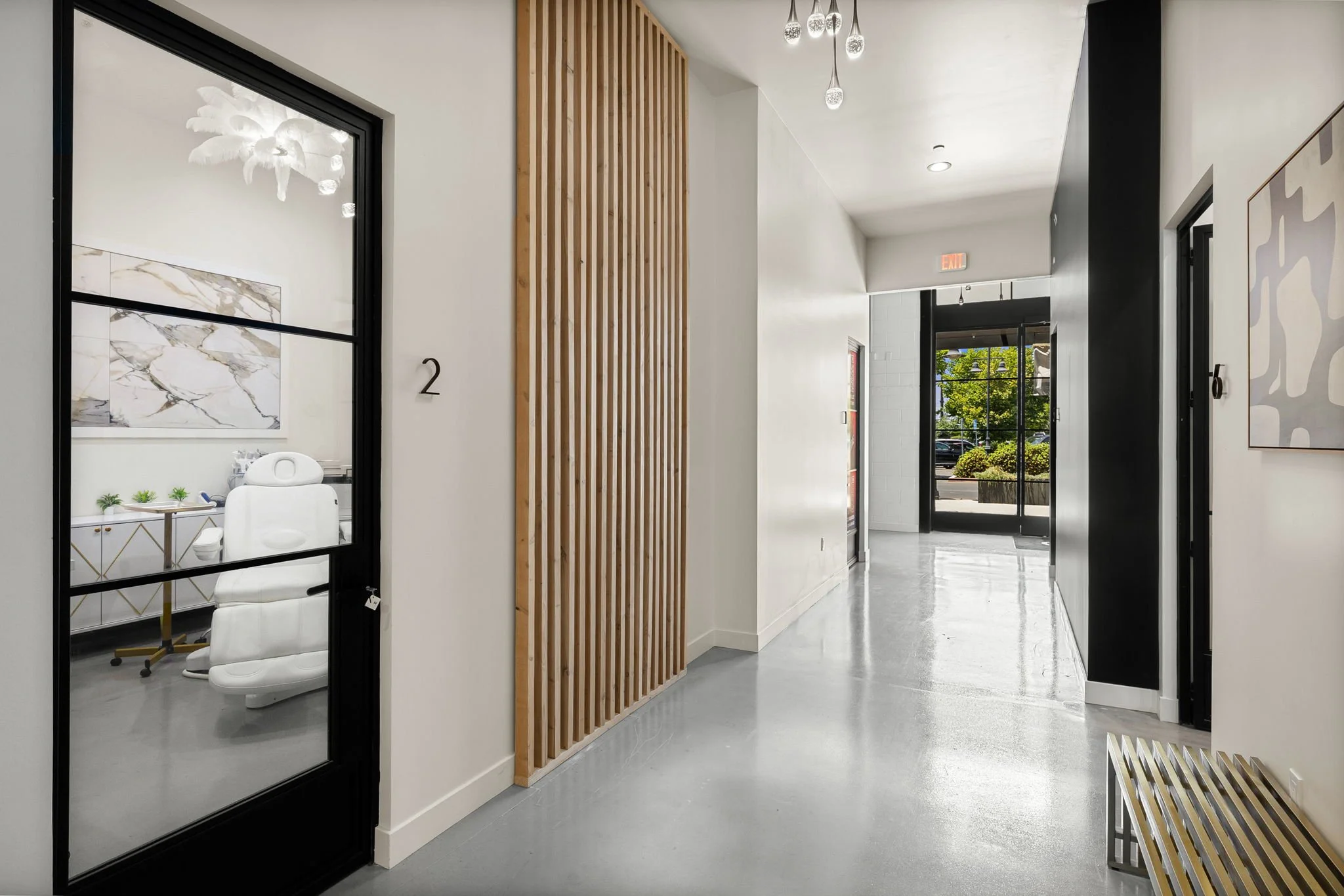 Modern office waiting area with glass door leading to a room with a white chair andside table, minimalist decor, light-colored floors, wooden slat wall, and large window showing outdoor greenery.