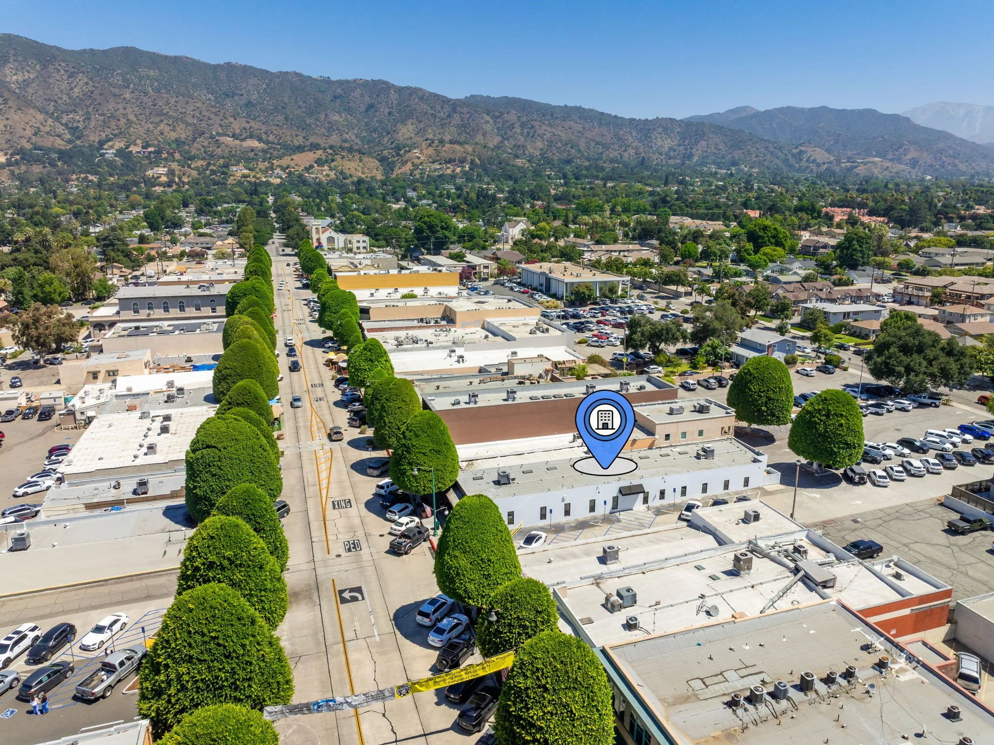 Aerial view of a commercial area with white buildings, parking lots, and green trees lining a central street, with mountains in the background.