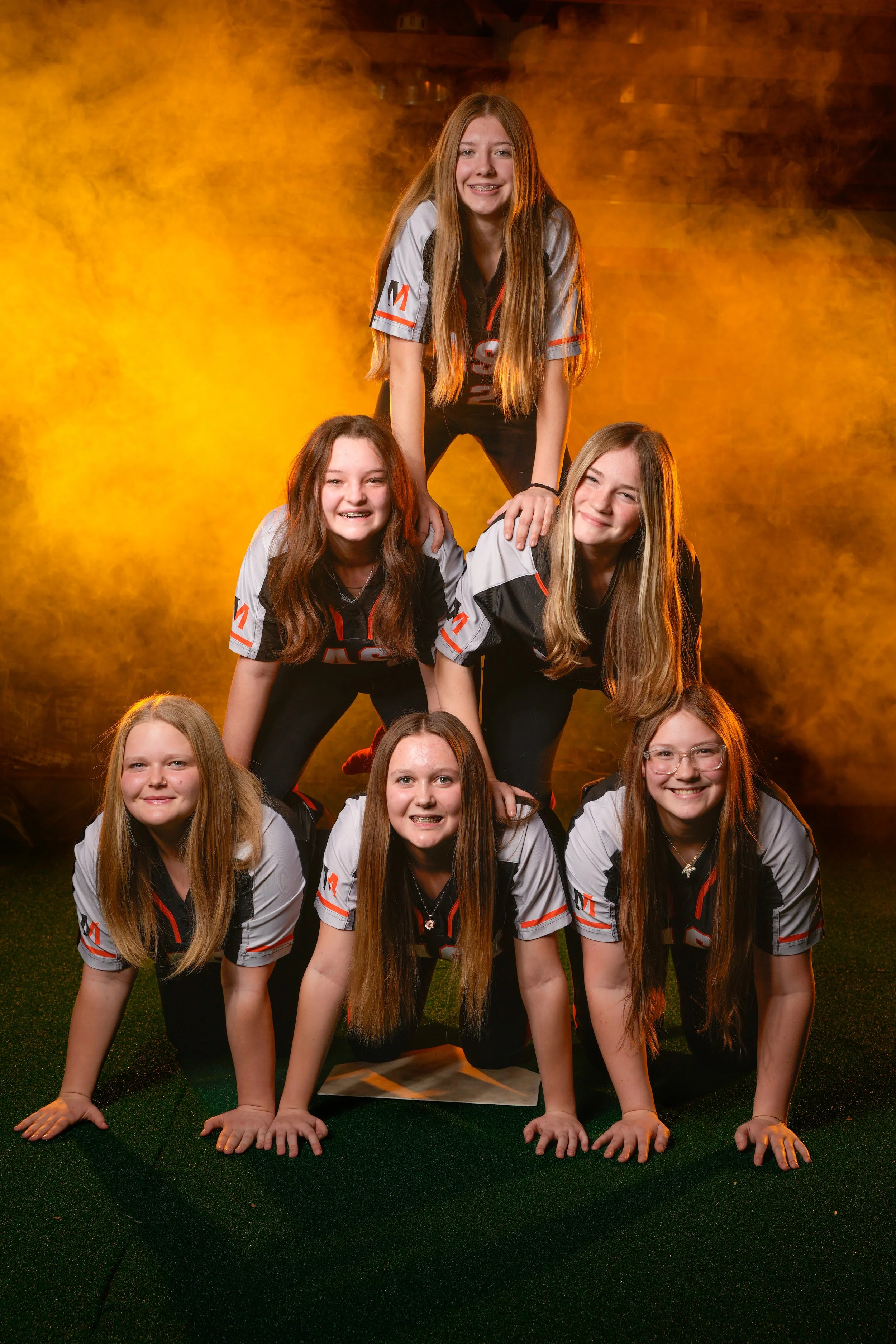 Seven young girls in matching sports uniforms forming a human pyramid, with three girls on the bottom, two in the middle, and one at the top, all smiling in front of a yellow-orange smoky background.