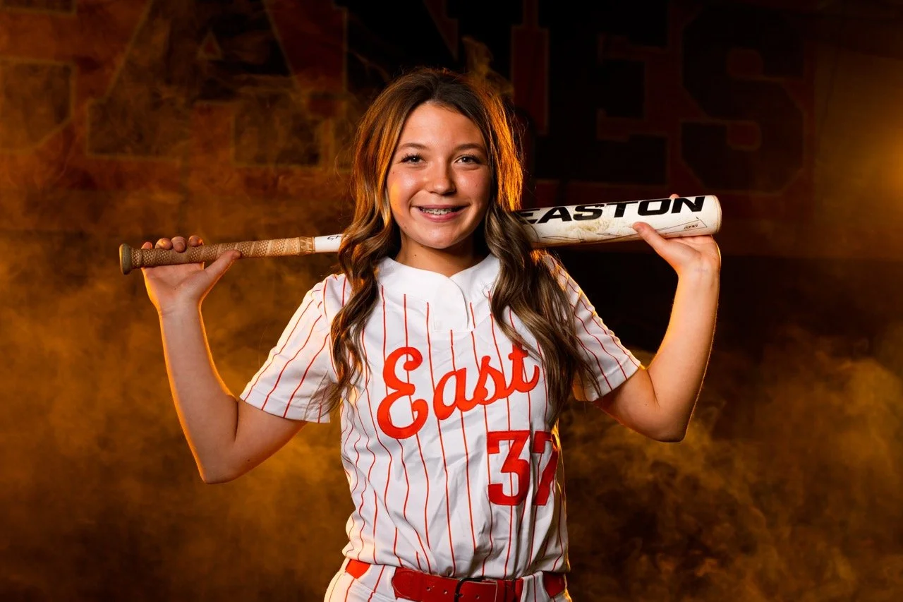 A young girl wearing a baseball jersey with 'East' and the number 37, smiling and holding a baseball bat across her shoulders.