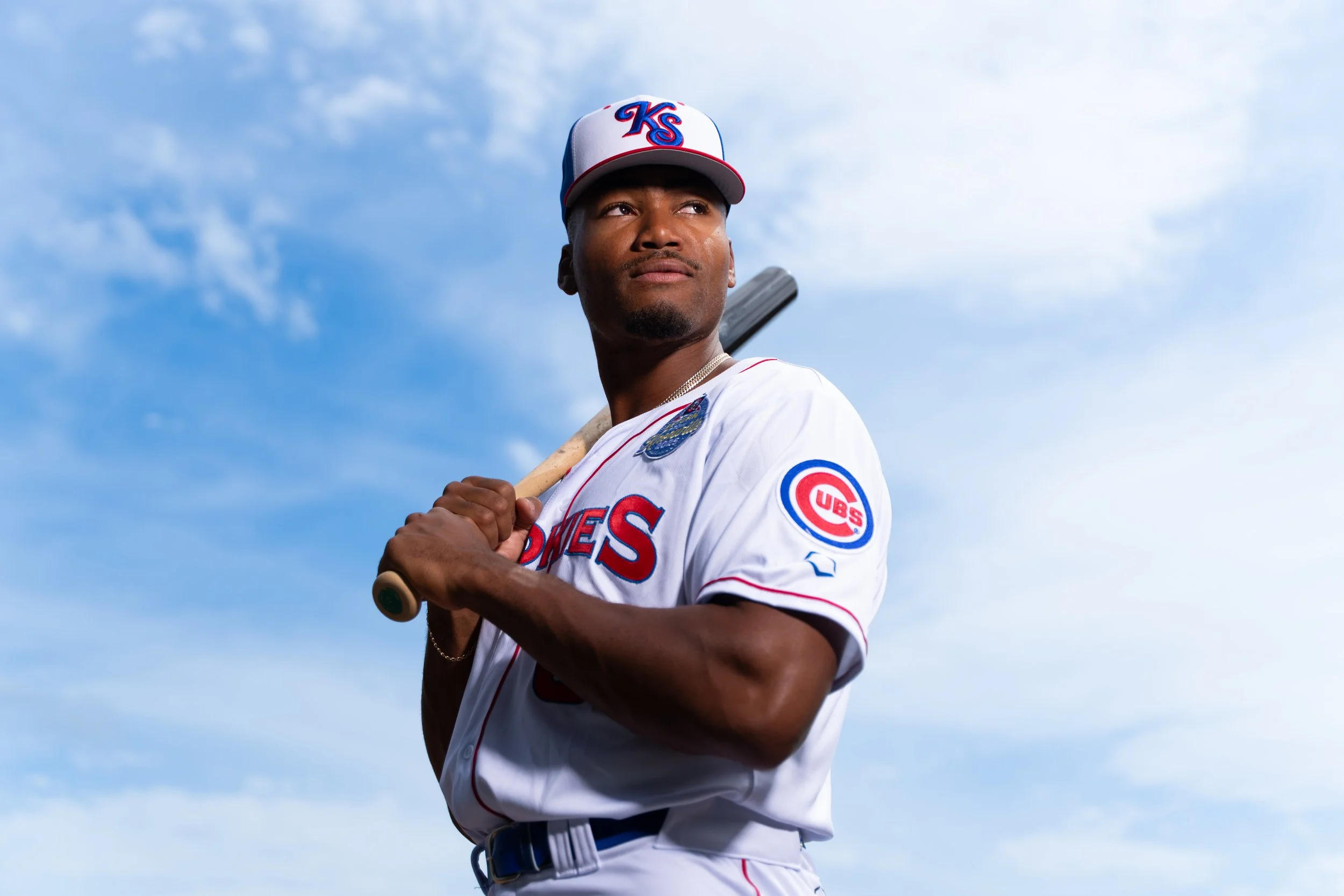 A baseball player in a Chicago Cubs uniform, holding a bat over his shoulder, standing outdoors under a partly cloudy sky.