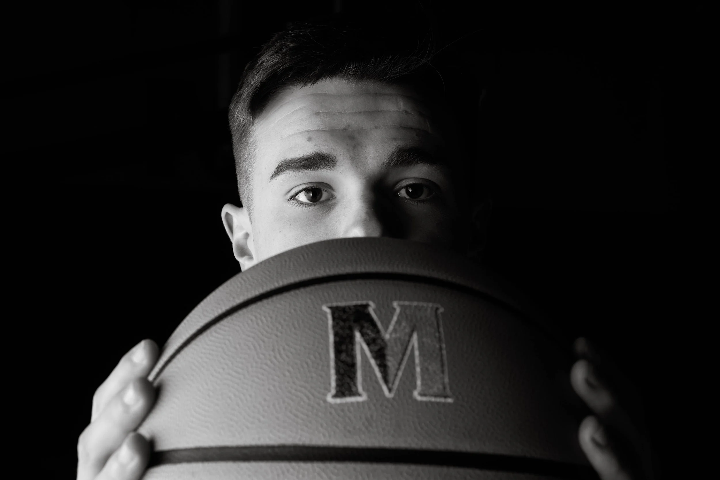 A young man with short hair holding a basketball in front of his face, with only his eyes and part of his forehead visible, against a dark background.