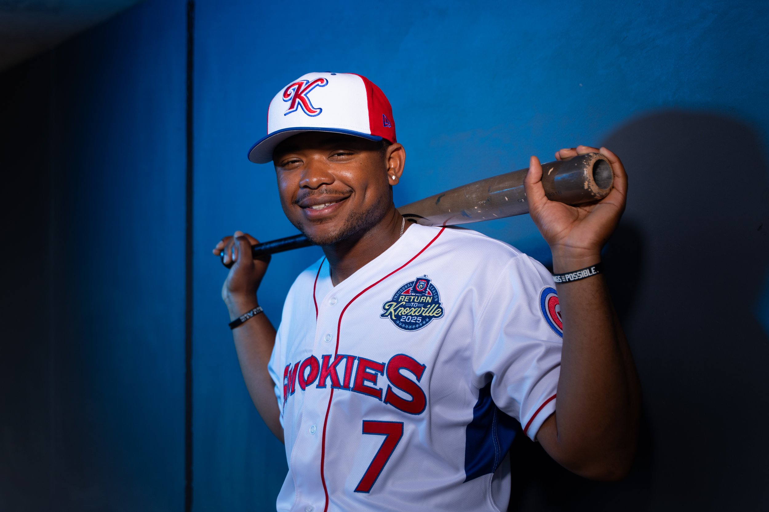 Smiling baseball player wearing a white jersey with red and blue accents, cap with a large 'K' logo, and holding a baseball bat over his shoulder against a dark background.