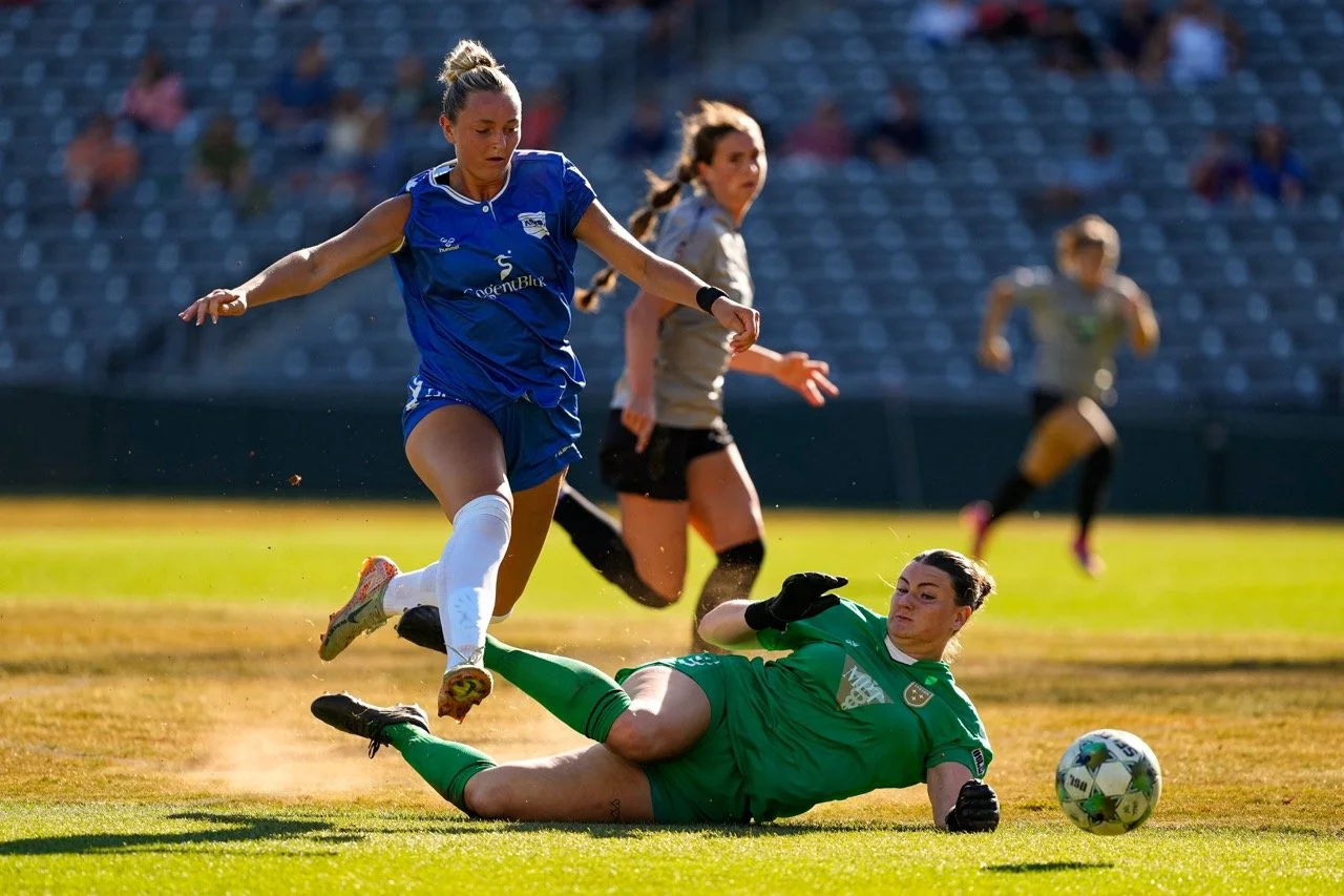 A female soccer player in a blue jersey attempting to score, while the goalkeeper in a green uniform makes a save on the field with other players and spectators in the background.