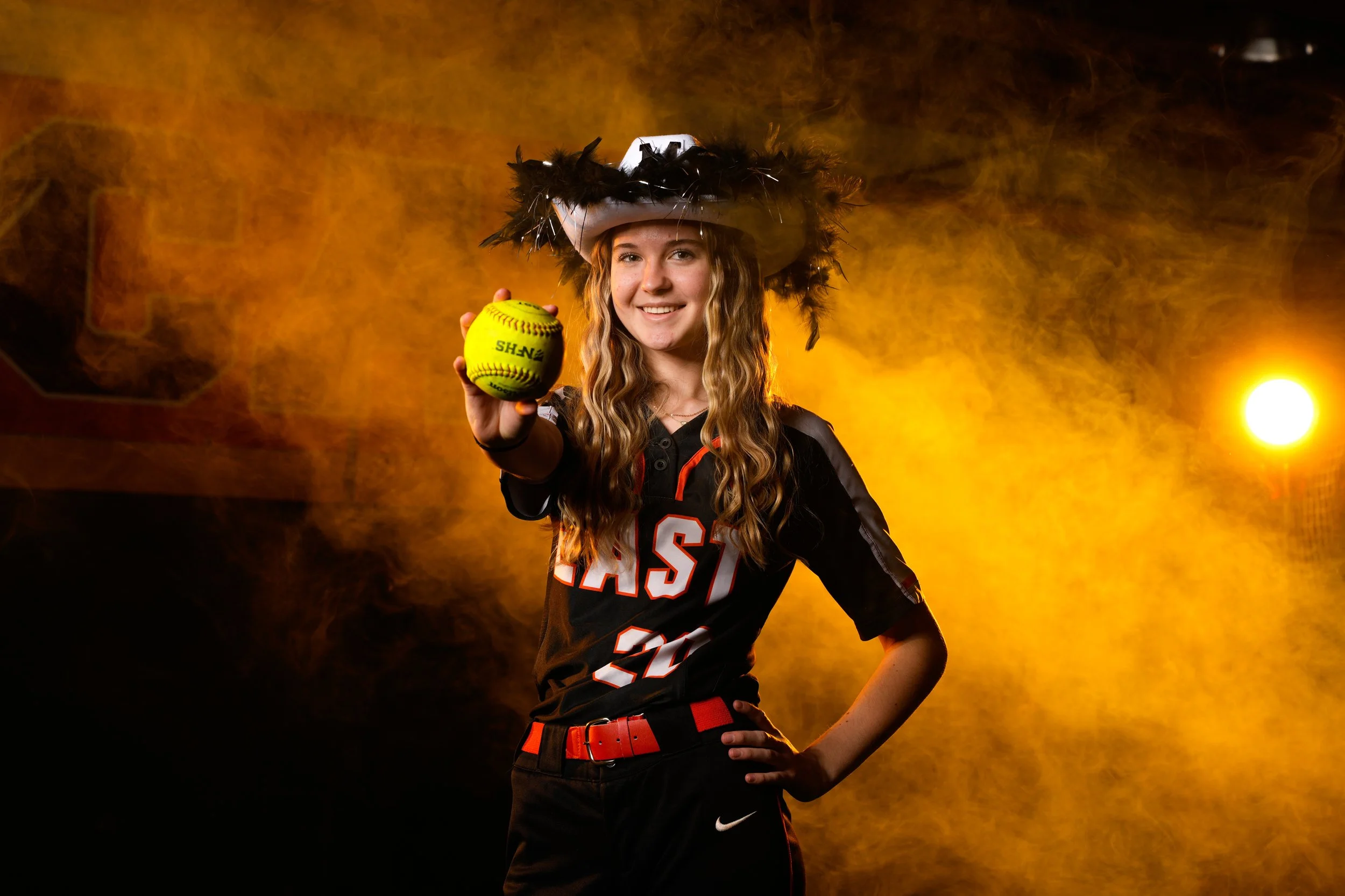 Young woman dressed as a softball player, wearing a black jersey with red and white accents and a red belt, holding a softball towards the camera, with a large hat decorated with black feathers, in front of an orange and yellow smoky background with 