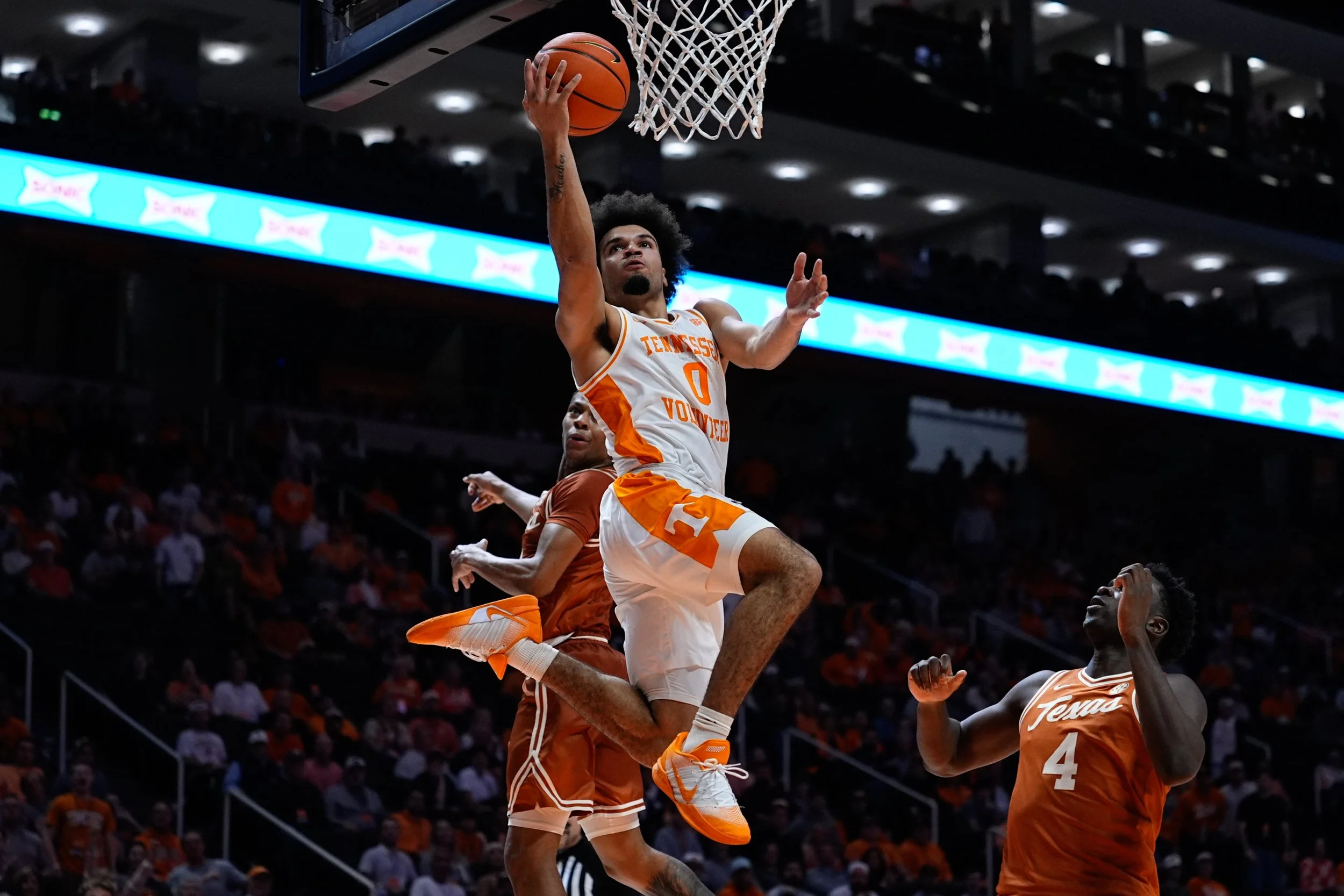 A basketball player from Tennessee Volunteers in white and orange uniform is dunking the basketball into the hoop, with two opponents in orange uniforms and one teammate watching from below, during a game in an arena filled with spectators.