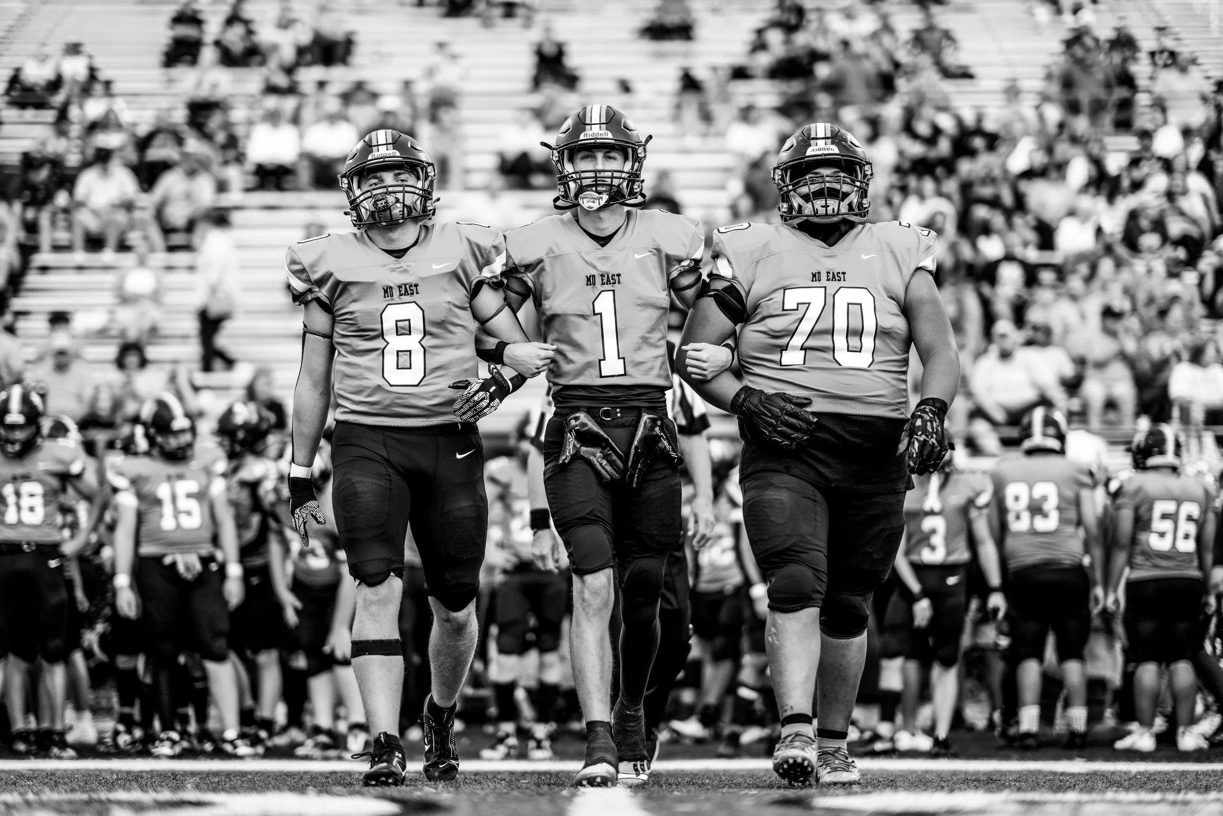 Three football players in uniform walking together on the field, with a crowd of spectators in the stands behind them.