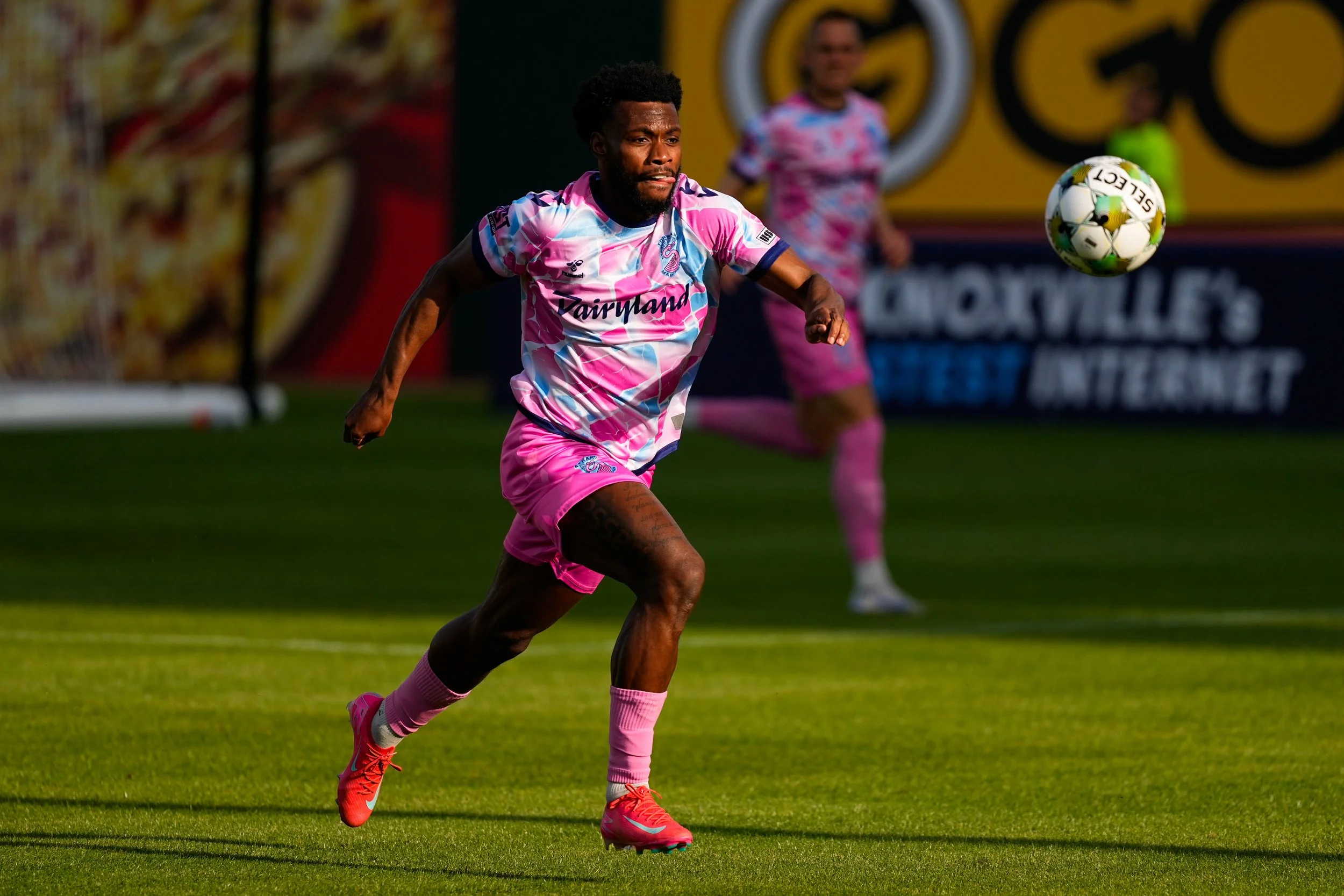 Soccer player in pink uniform running on field with a soccer ball in mid-air