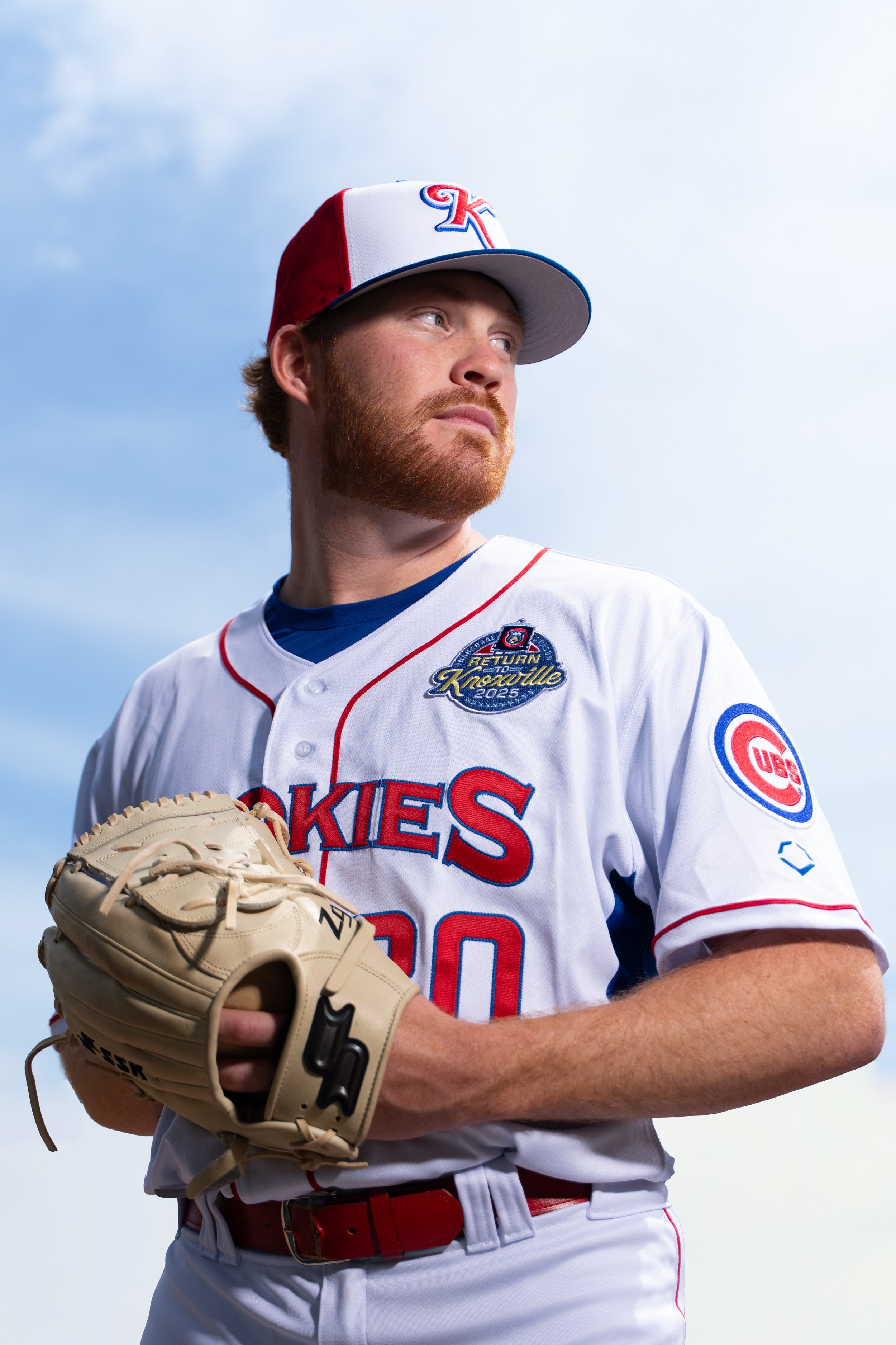 A male baseball player in a white, red, and blue uniform holding a tan baseball glove, wearing a cap with a red, white, and blue logo, standing against a cloudy sky.