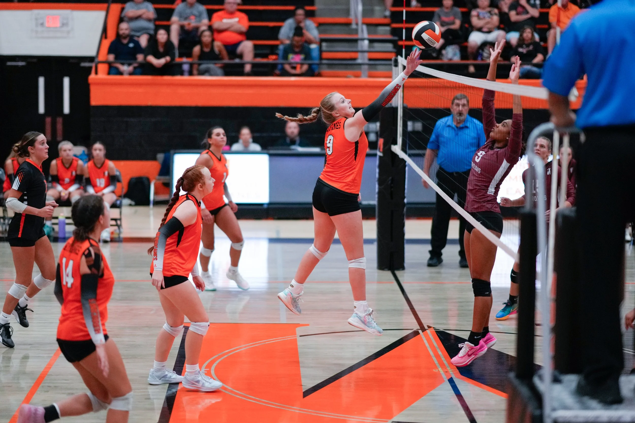 A volleyball game where a player in an orange uniform is jumping to hit the ball at the net, while a player in a maroon uniform is attempting to block. Other players are positioned on the court, and spectators can be seen watching from the stands.