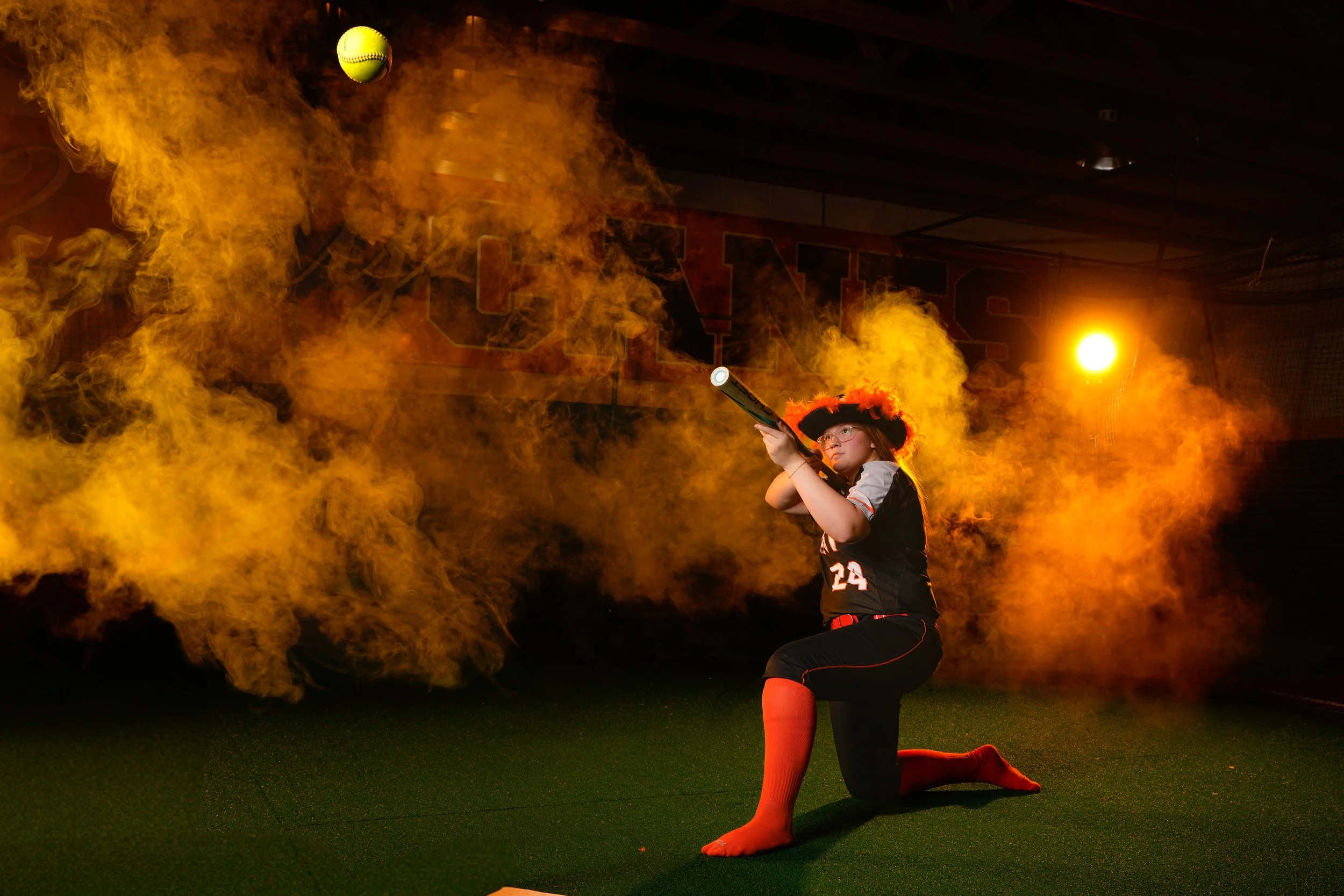 A girl dressed in a black and red sports uniform, with orange socks, is kneeling on one knee on a green surface. She is aiming a gun with a flashlight attached, with a smoke or fog surrounding her. The background is dark with intense warm lighting, i