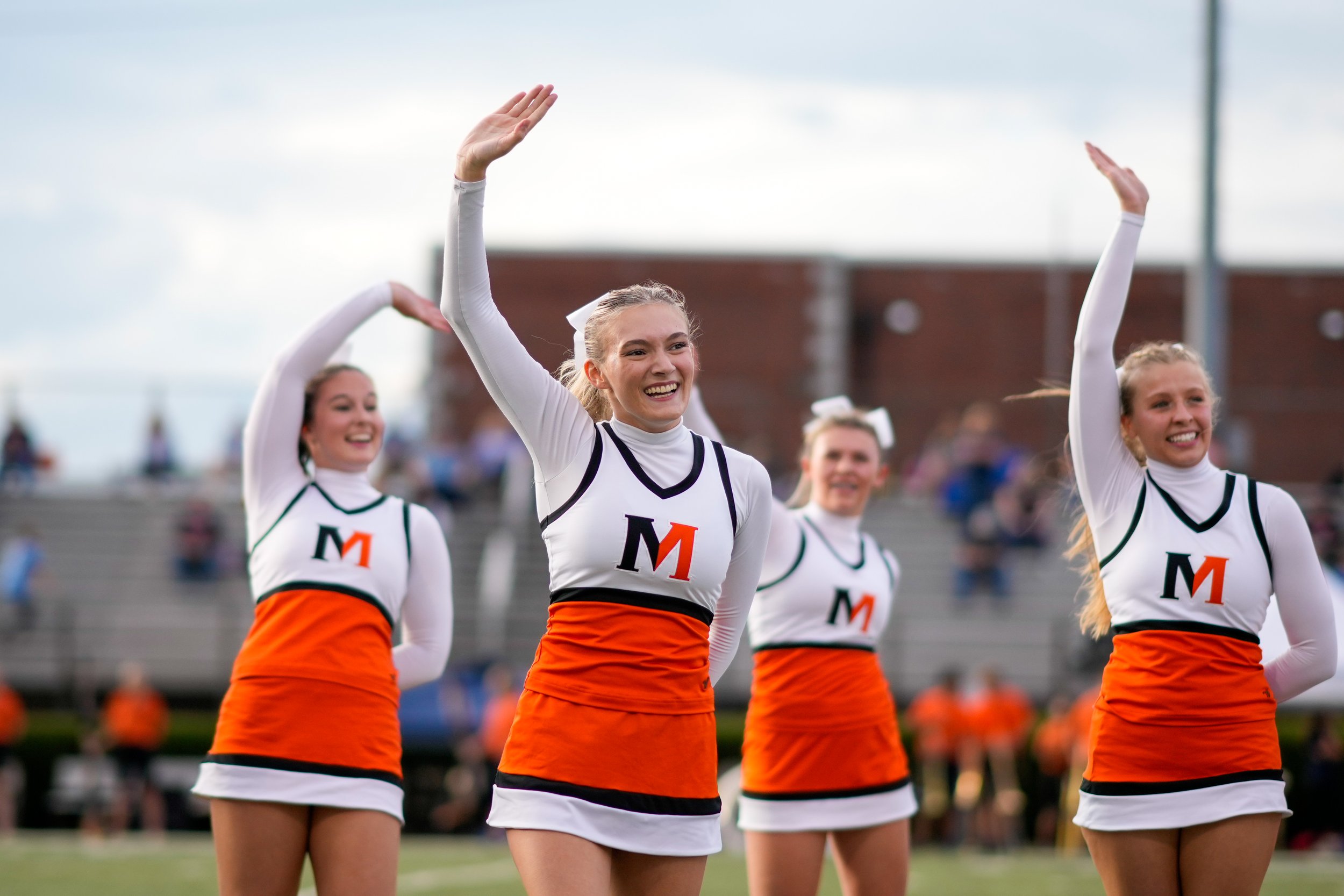 Cheerleaders in orange and white uniforms performing a cheer routine on a football field, with fans in the background.