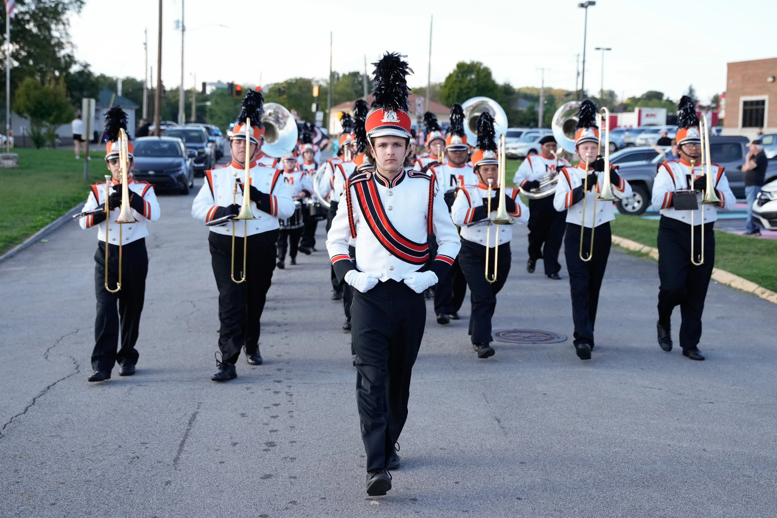 A marching band dressed in white and black uniforms with red and black stripes, wearing tall black feathered hats, performing in a parade on a street with cars and trees in the background.