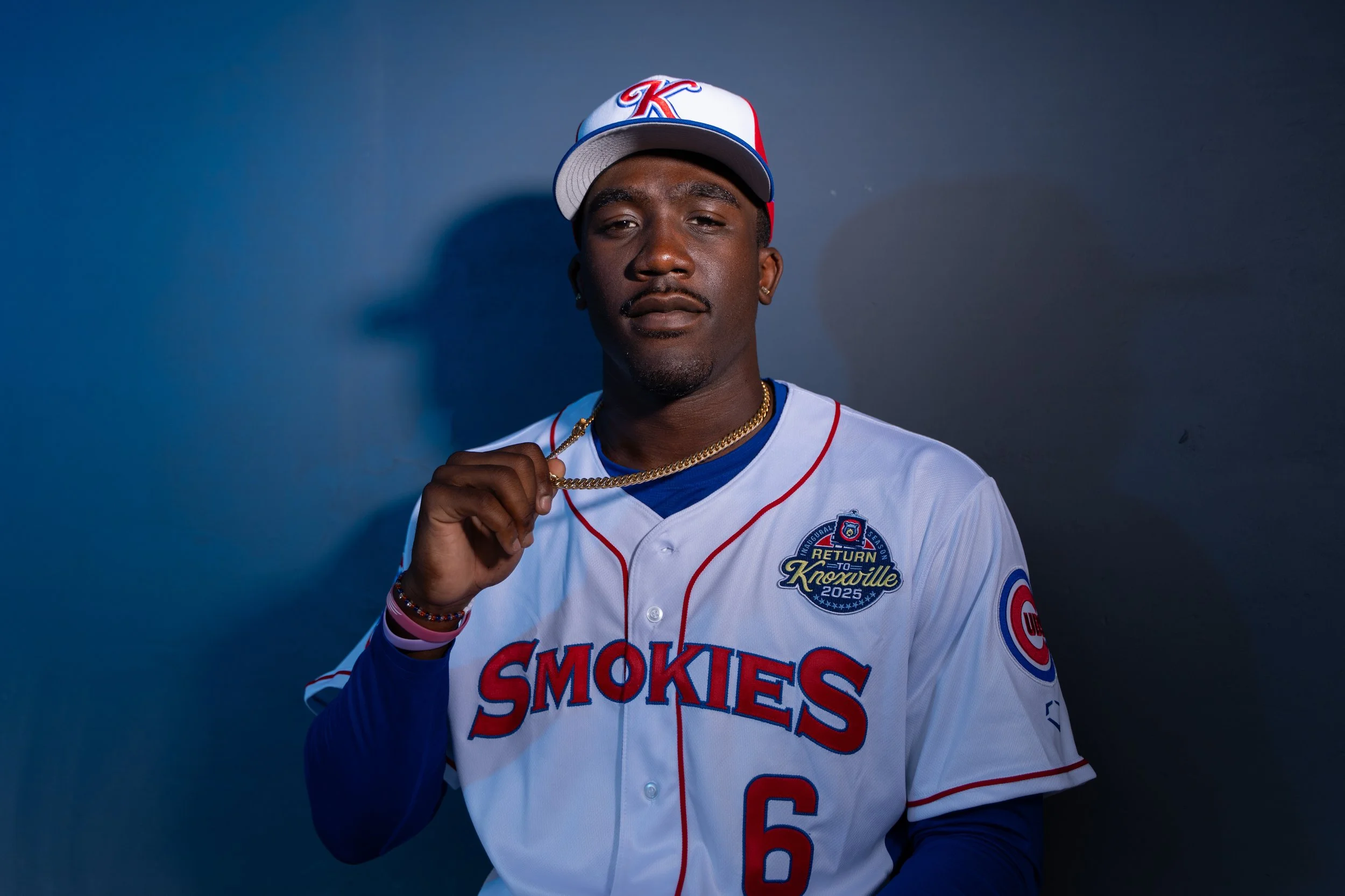 Young man wearing a white and red baseball jersey with 'Smokies' written on it and the number 6, a matching cap with red, white, and blue, holding a gold chain, standing against a dark background.