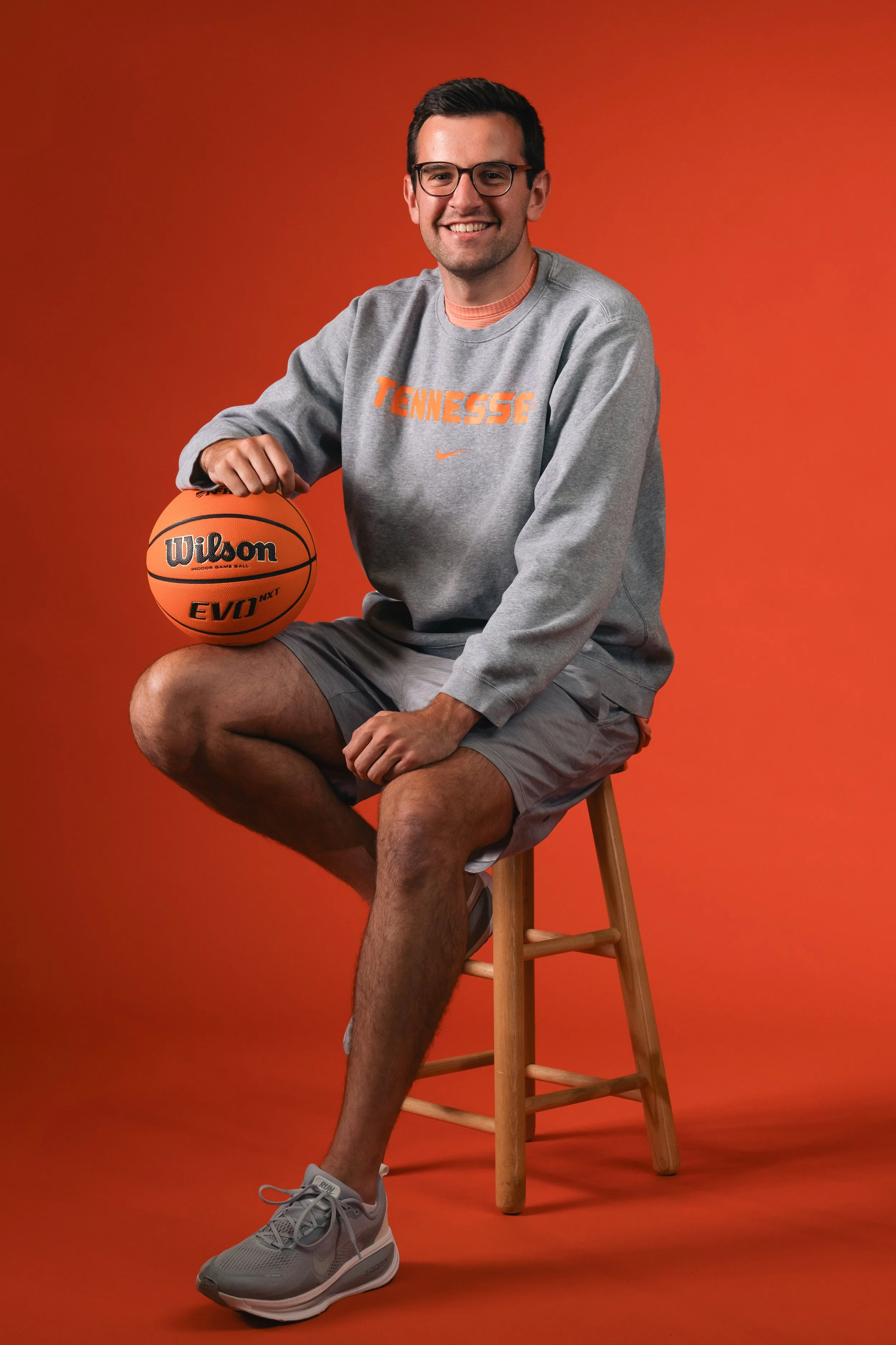 A young man with glasses, wearing a gray Tennessee sweatshirt and shorts, sitting on a wooden stool with a basketball in his right hand, smiling at the camera against an orange background.