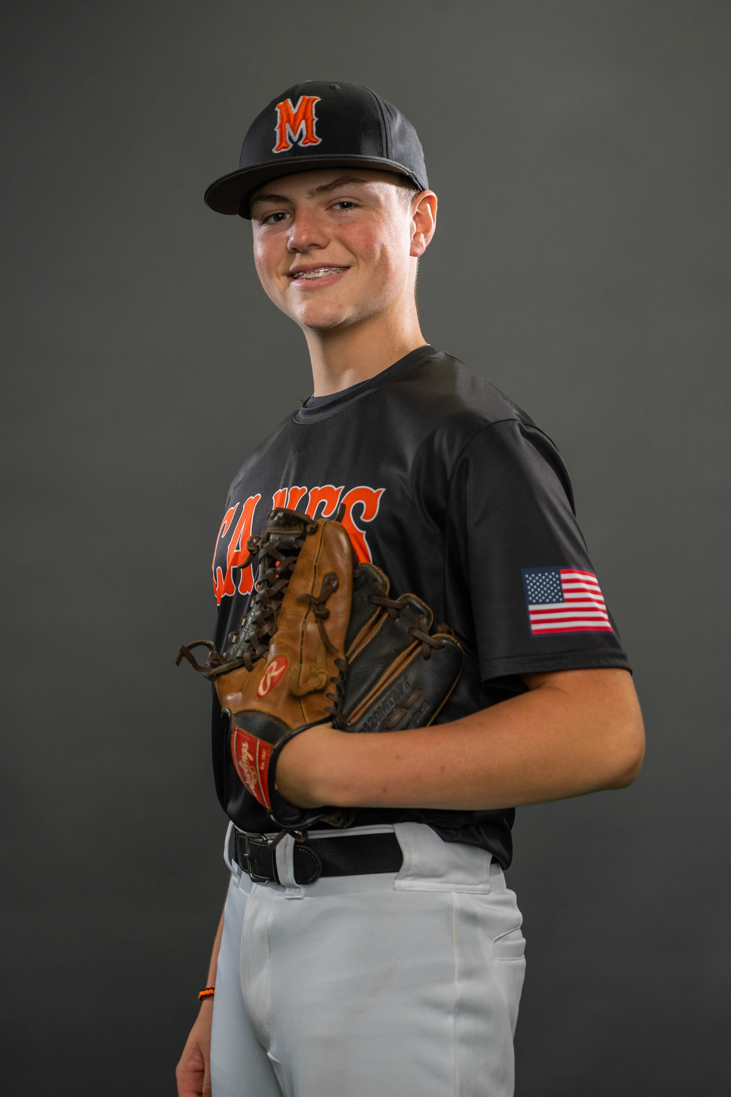 A young baseball player in uniform holding a glove, smiling, with an American flag patch on his sleeve, against a gray background.