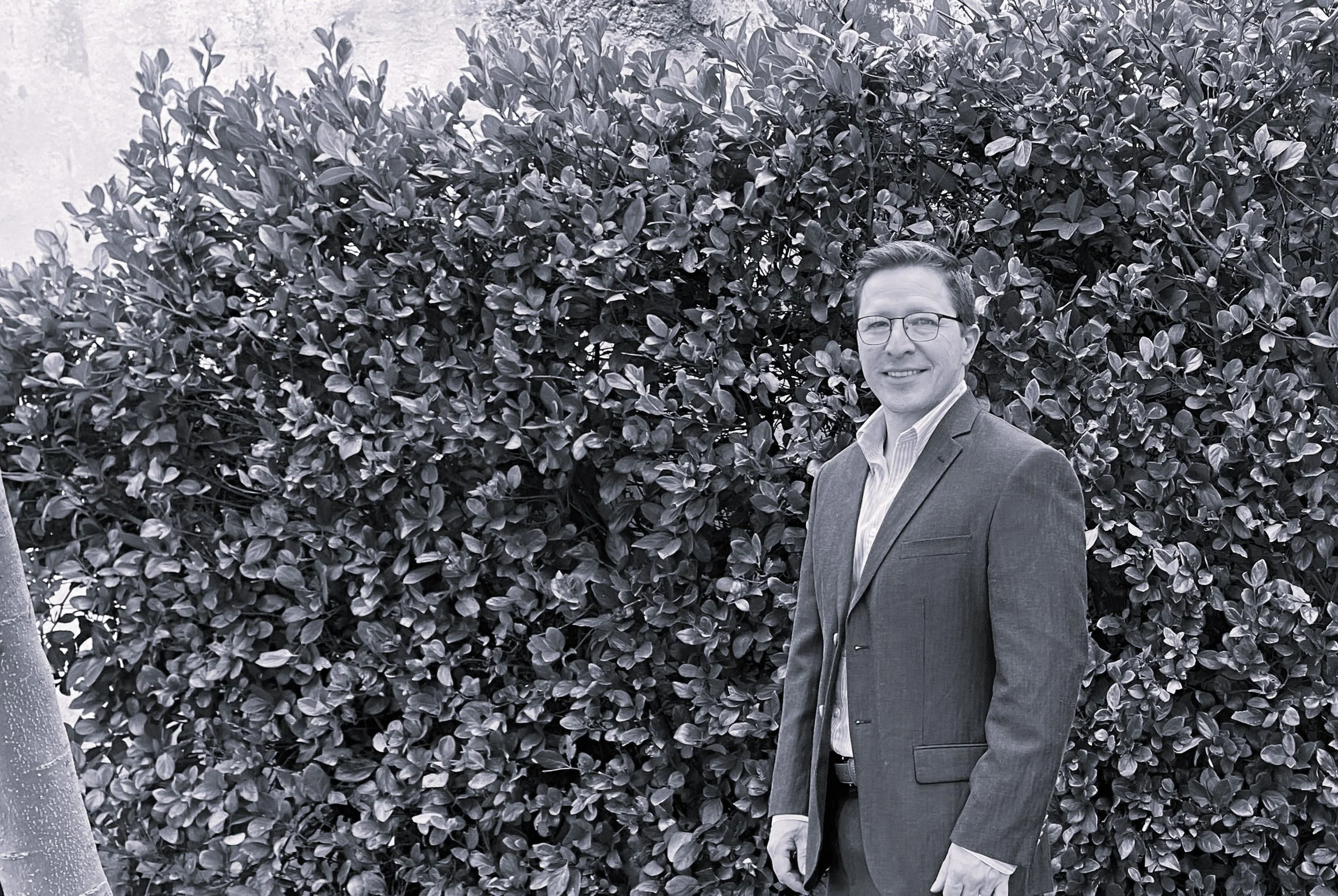A man dressed in a suit and glasses standing in front of a large bush or hedge.