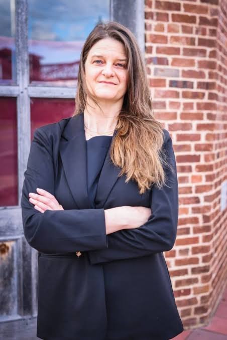 Woman in professional attire stands in front of brick wall.