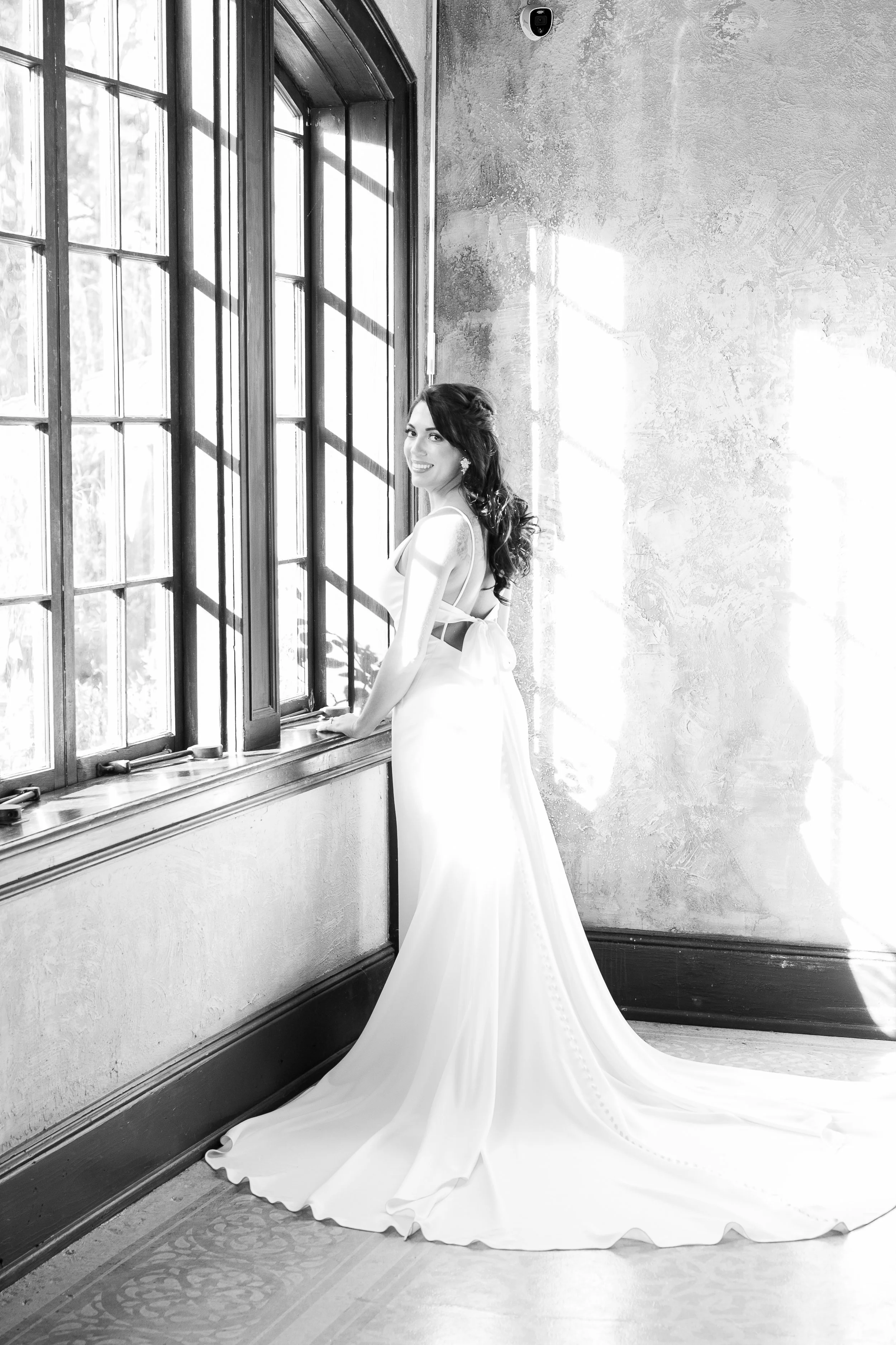Black and white photo of a smiling woman in a wedding dress standing by a large window, looking outside.